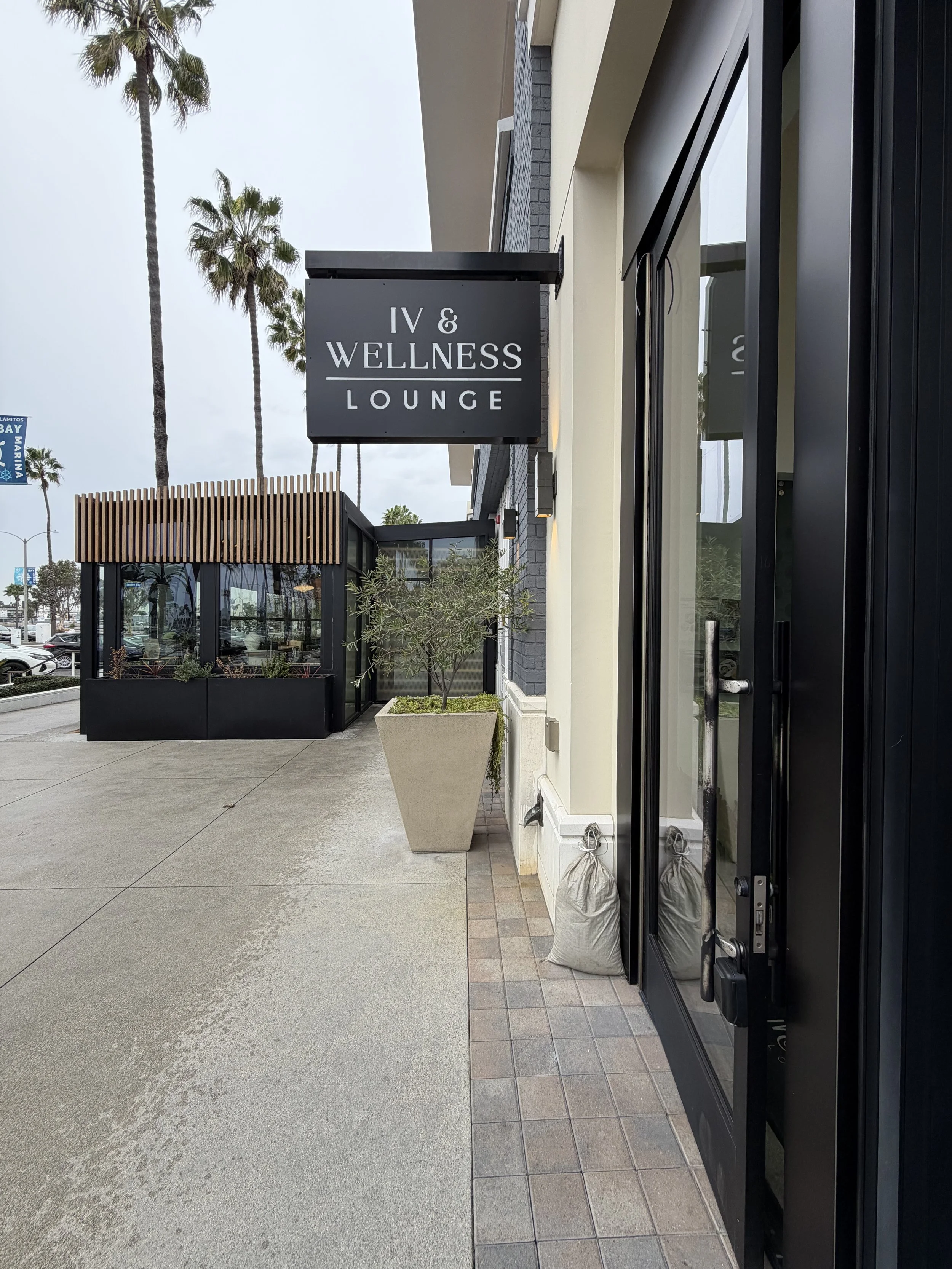Outside view of a wellness lounge with black sign reading 'IV & Wellness Lounge', large glass doors, potted plant, sidewalk, and palm trees in the background.