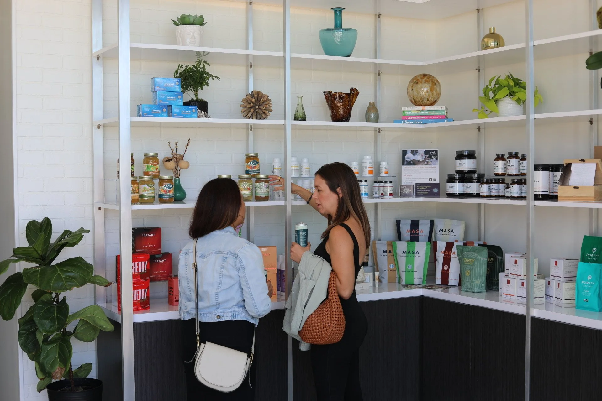 Two women shopping for health supplements in a store with white shelves filled with various jars, boxes, and packages of herbal and dietary products.