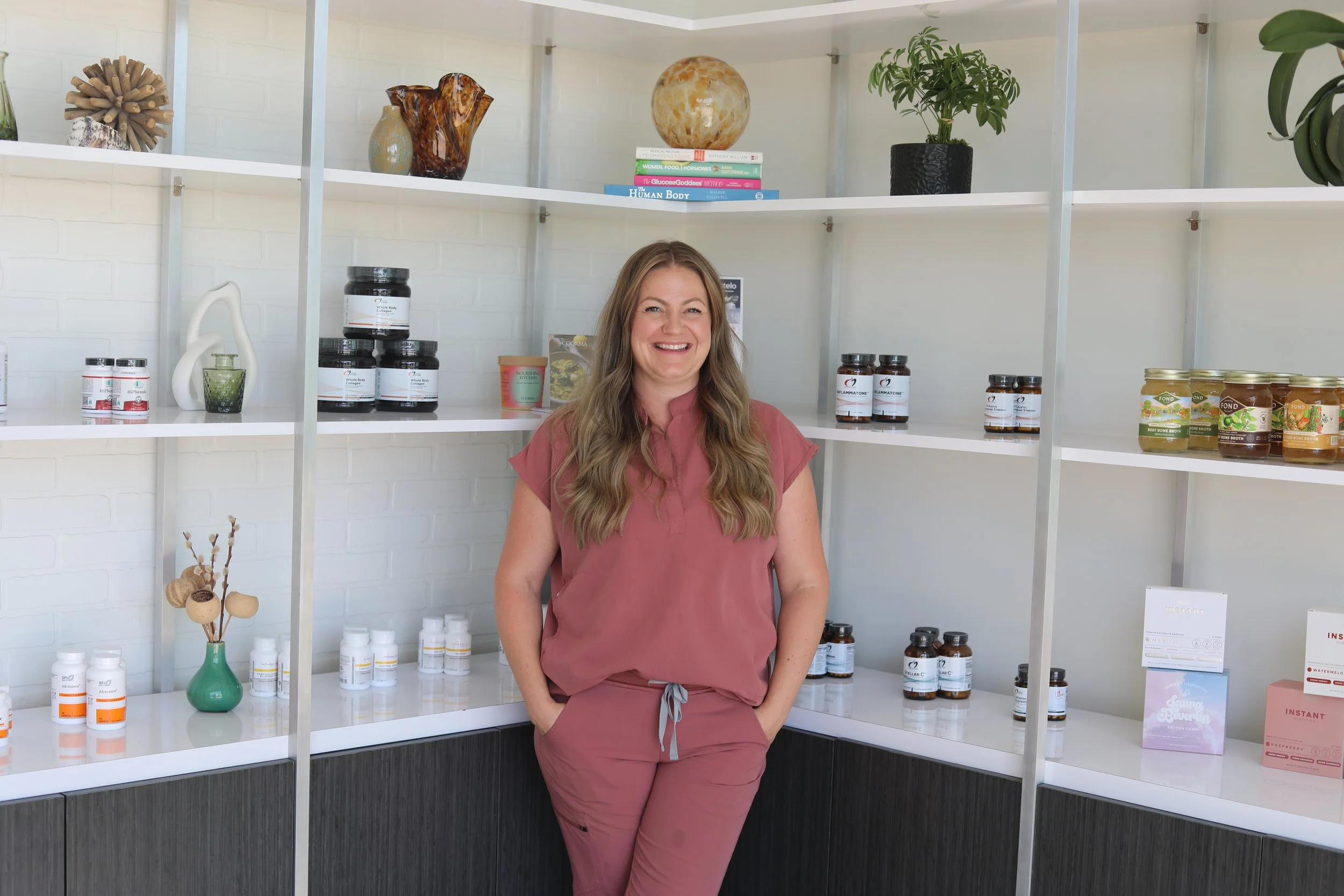 A woman with long wavy hair smiling and standing with her hands in her pockets in front of white shelves filled with containers, bottles, jars, decorative items, and books on a white brick wall background.
