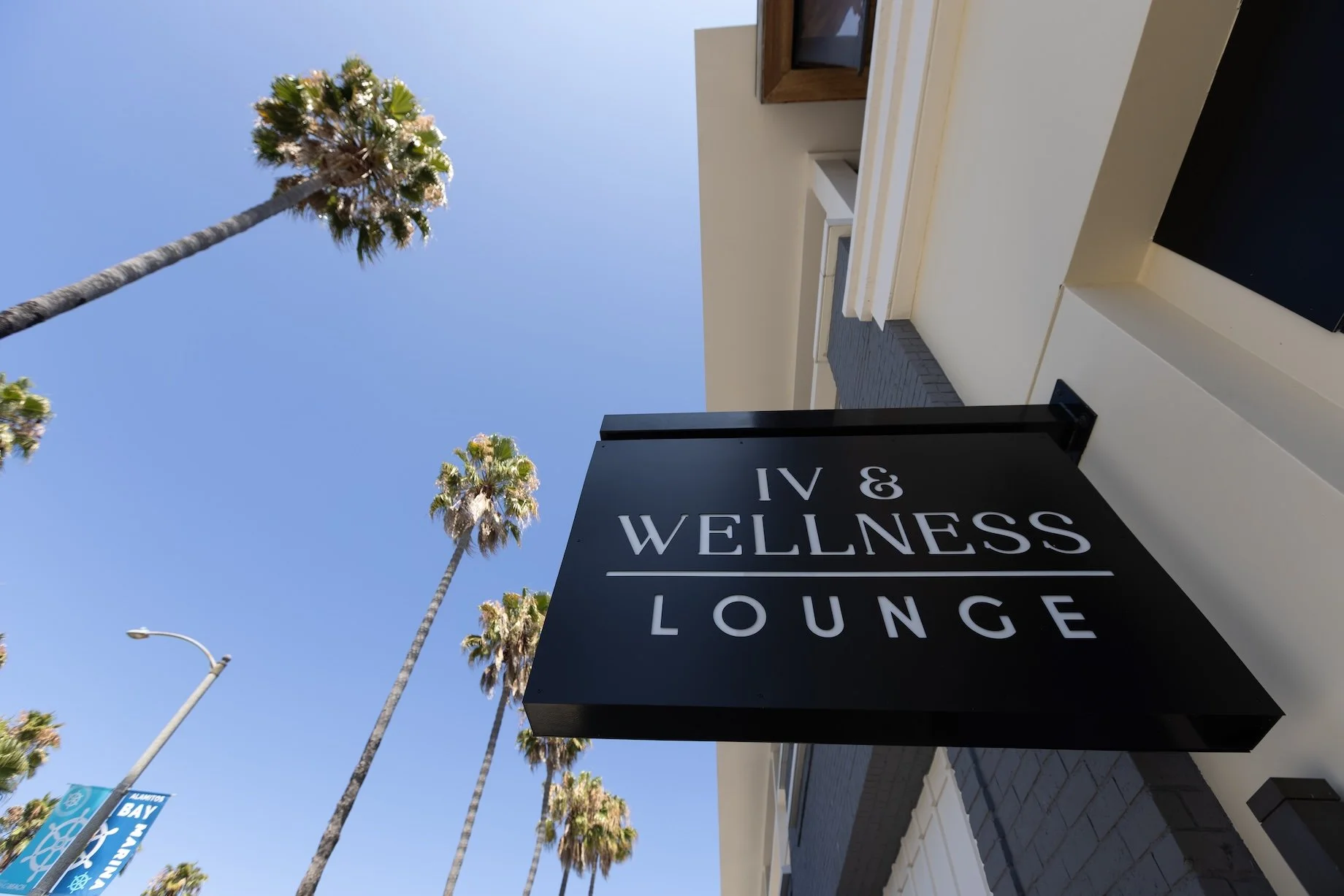 Black signboard with white text reading "IV & WELLNESS LOUNGE" mounted on a building exterior, with palm trees and a clear blue sky in the background.