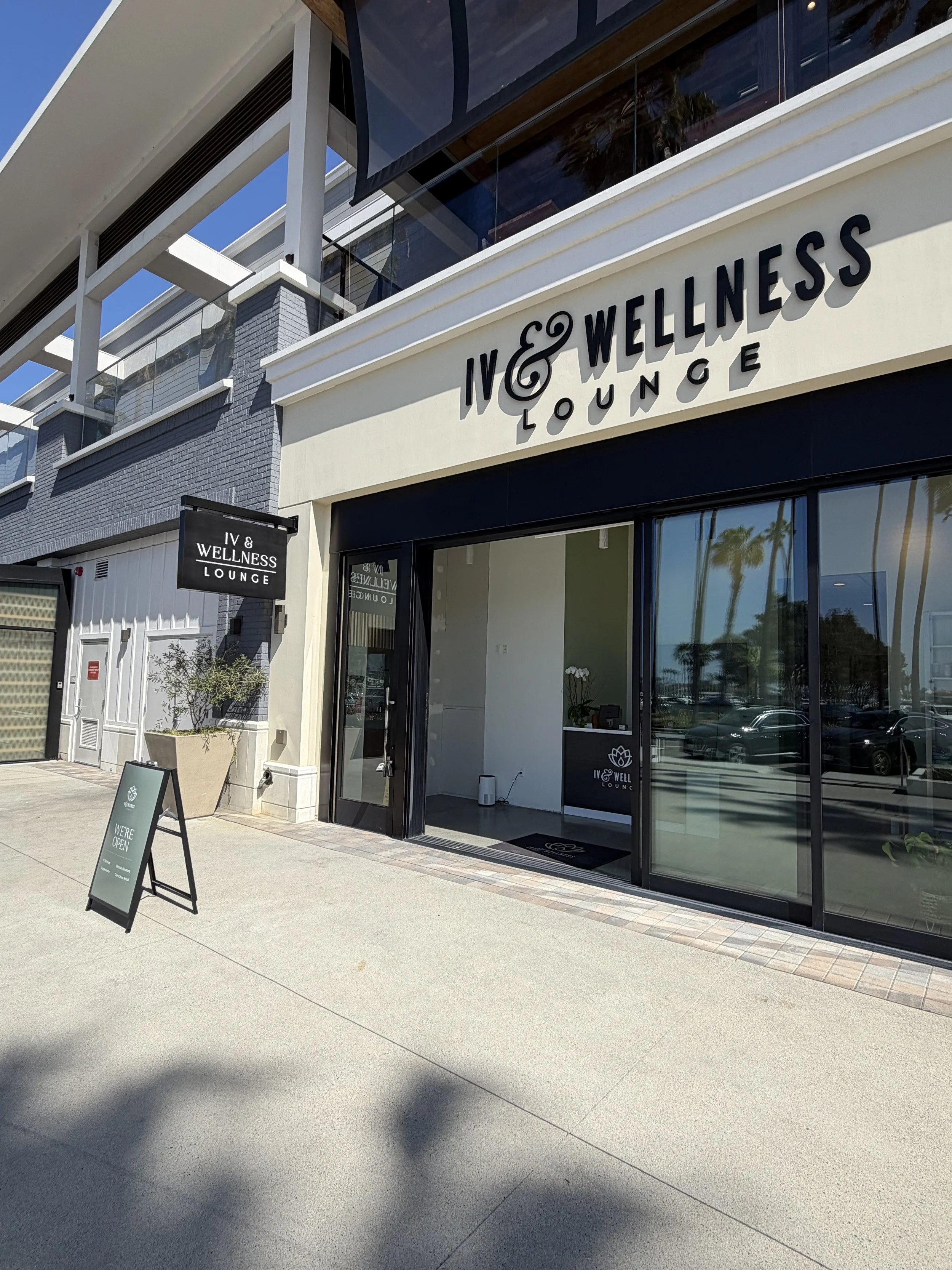 Exterior view of IV & Wellness Lounge with glass doors, a black and white sign, and large potted plant outside on a sunny day.