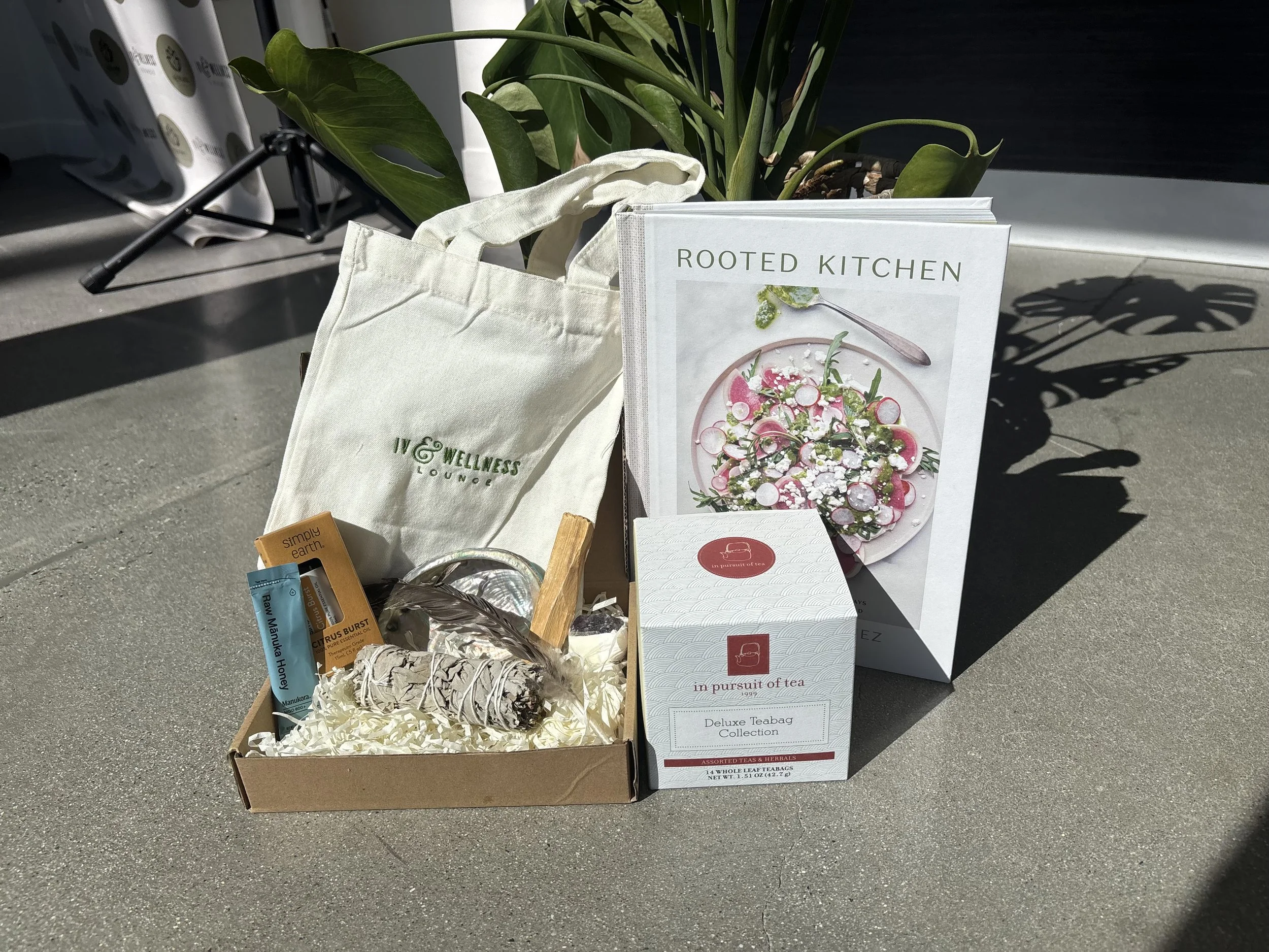 A wellness lounge gift bag, a cookbook titled 'Rooted Kitchen', and a tea collection box are displayed outdoors on a gray surface with a leafy plant in the background.