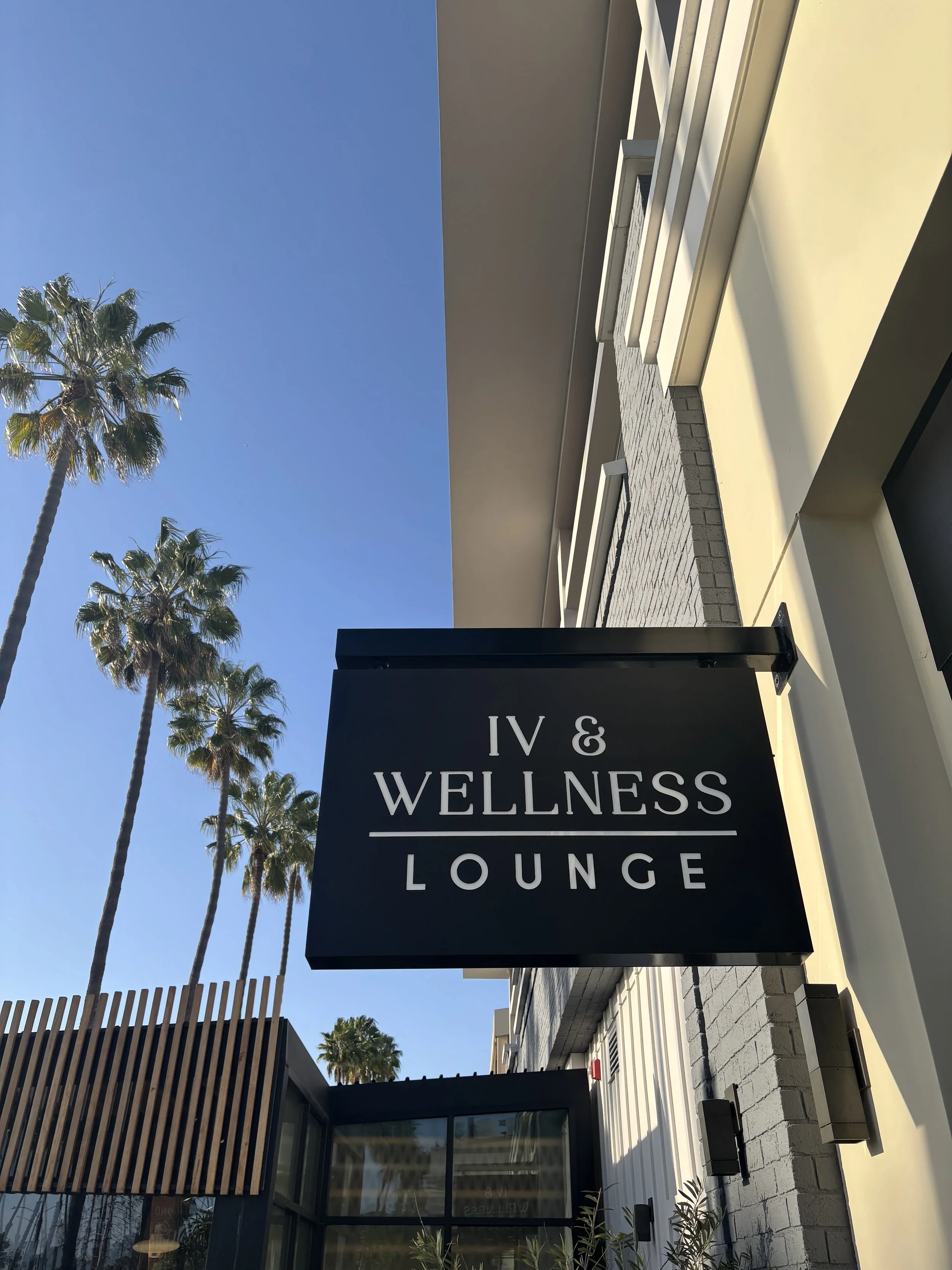 A black sign with white text reading 'IV & WELLNESS LOUNGE' hangs outside a building, with palm trees and a clear blue sky in the background.