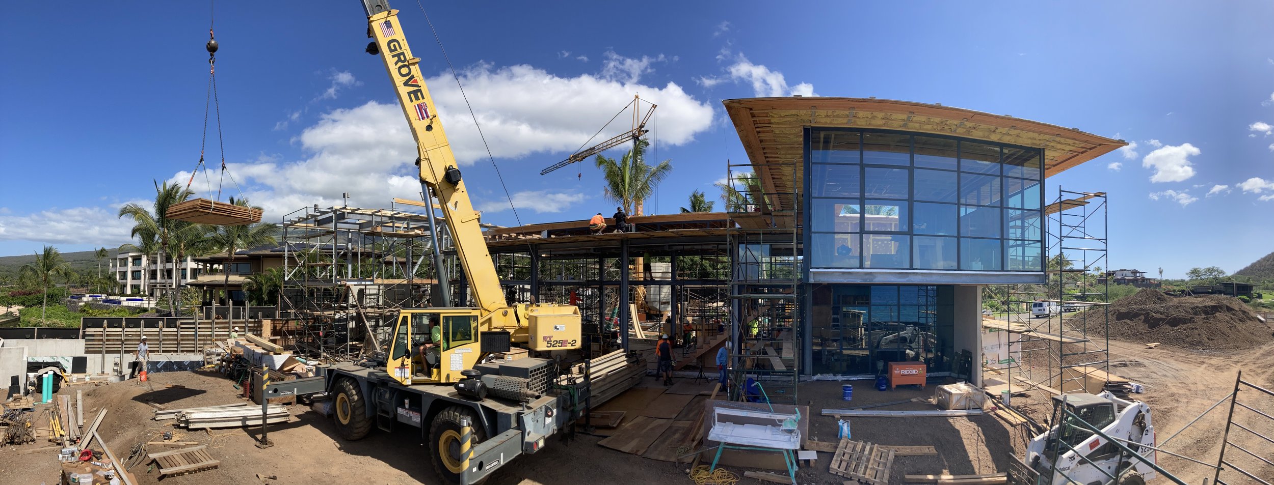 Construction site with building under construction, a crane lifting materials, scaffolding, workers, and a modern glass building with a curved wooden roof.
