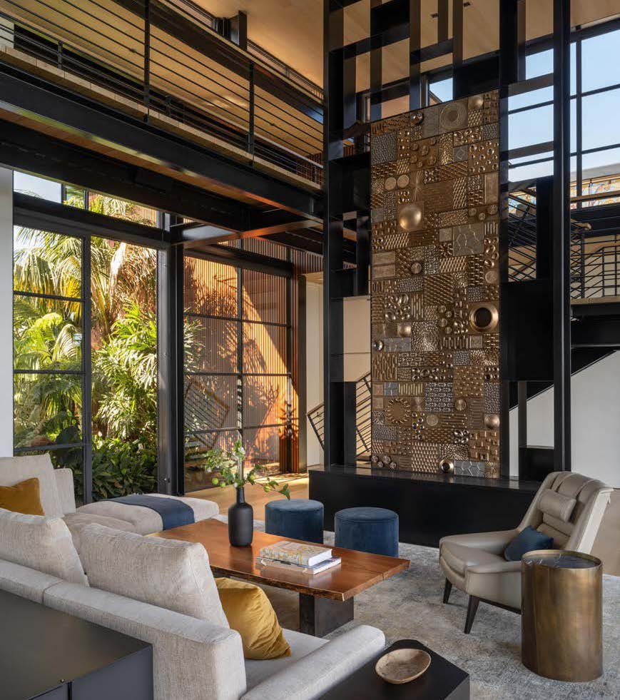 Modern living room with large glass windows, a variety of beige, white, and blue seating, a wooden coffee table, decorative vases, an ornate wall sculpture, and lush green outdoor plants visible through the windows.