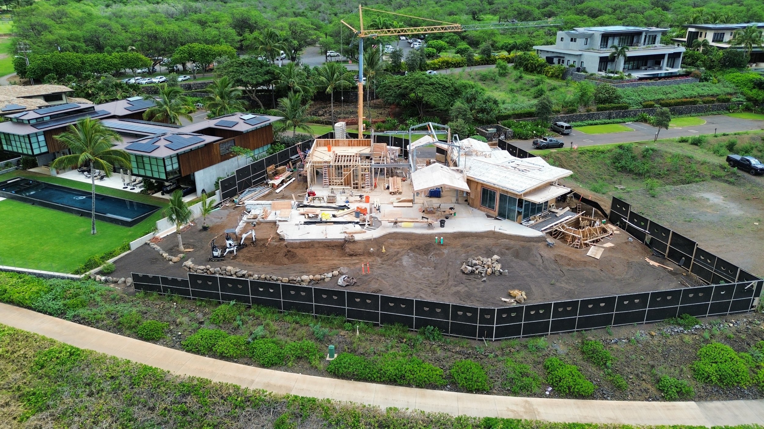 Aerial view of a house under construction with construction equipment, wooden framing, and construction materials, surrounded by a black fence, green landscape, and neighboring modern houses.