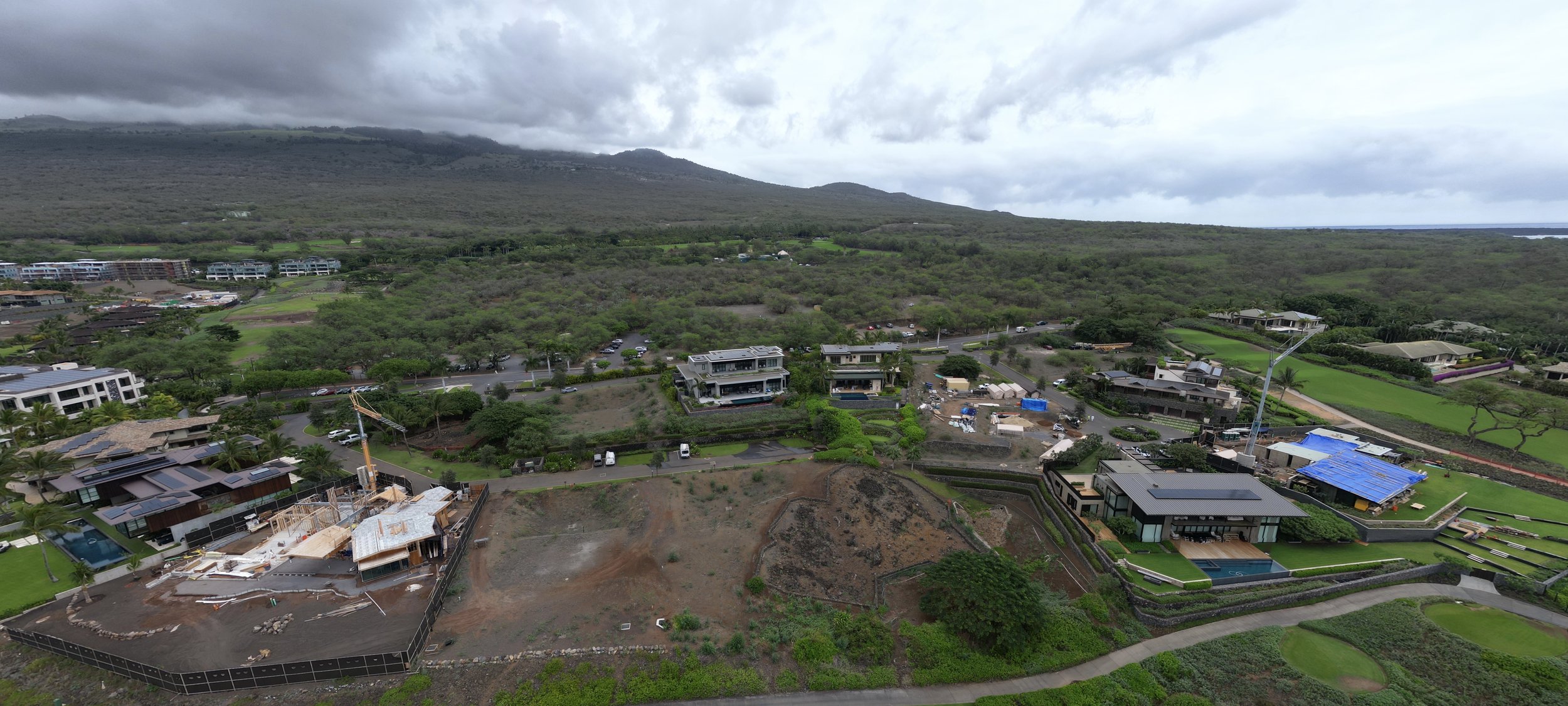 Aerial view of a suburban neighborhood with houses, construction, greenery, and mountains in the background under cloudy skies.