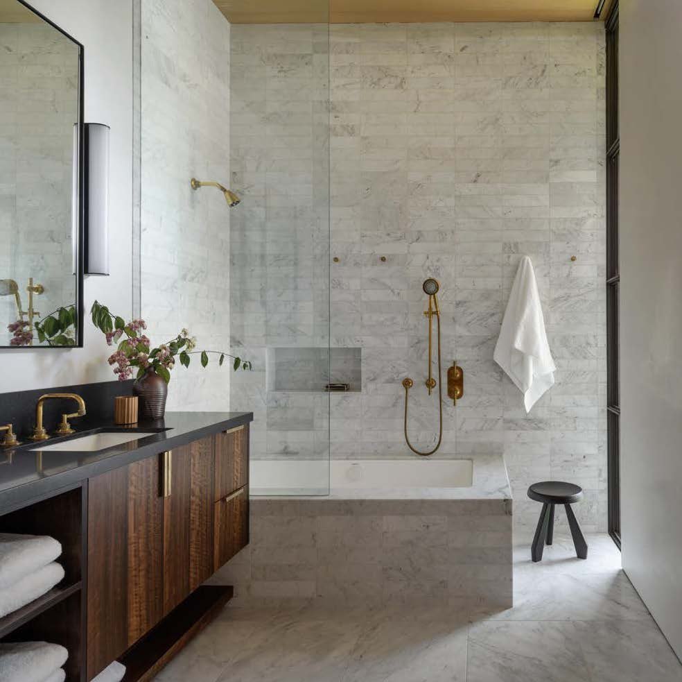 Modern bathroom with marble walls and floor, featuring a walk-in shower with gold fixtures, a dark wooden vanity with a black countertop, a large mirror, and a small black stool near a tall window.