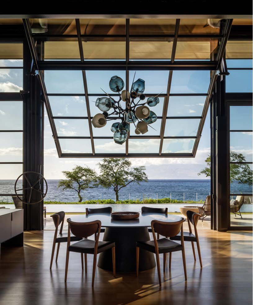 Modern dining room with a round black table and six wooden chairs, overlooks a scenic view of trees and the ocean through large glass sliding doors, a chandelier with multiple light bulbs hangs from the ceiling, and a small outdoor seating area is vi