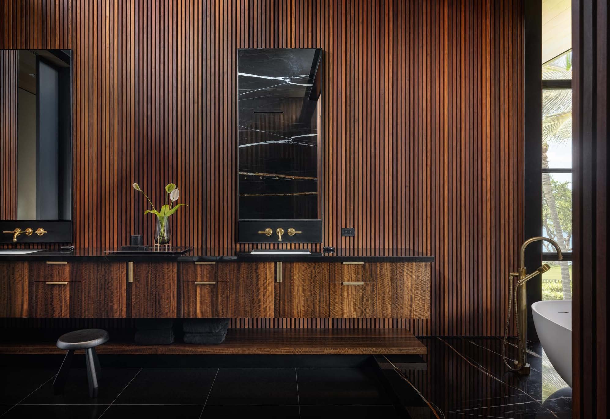 Bathroom with dark wood paneling, black marble vanity with gold fixtures, a mirror, a small stool, and a bathtub with a gold faucet near a window showing outdoor greenery.
