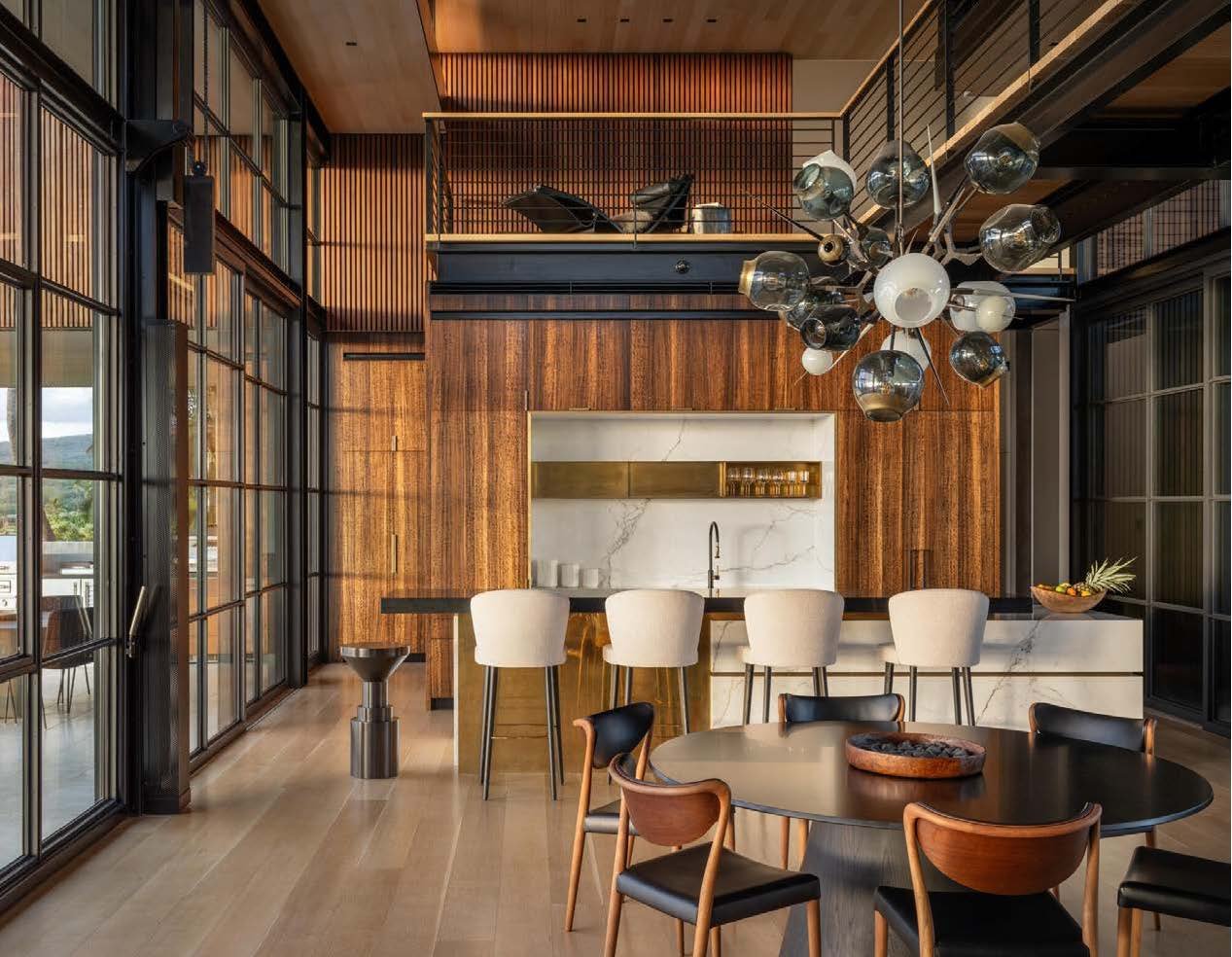 Modern kitchen and dining area with wood-paneled walls, a large marble backsplash, and a high ceilings. There is a black round table with chairs, a marble kitchen island with barstools, and a black modern chandelier with multiple glass bulbs.