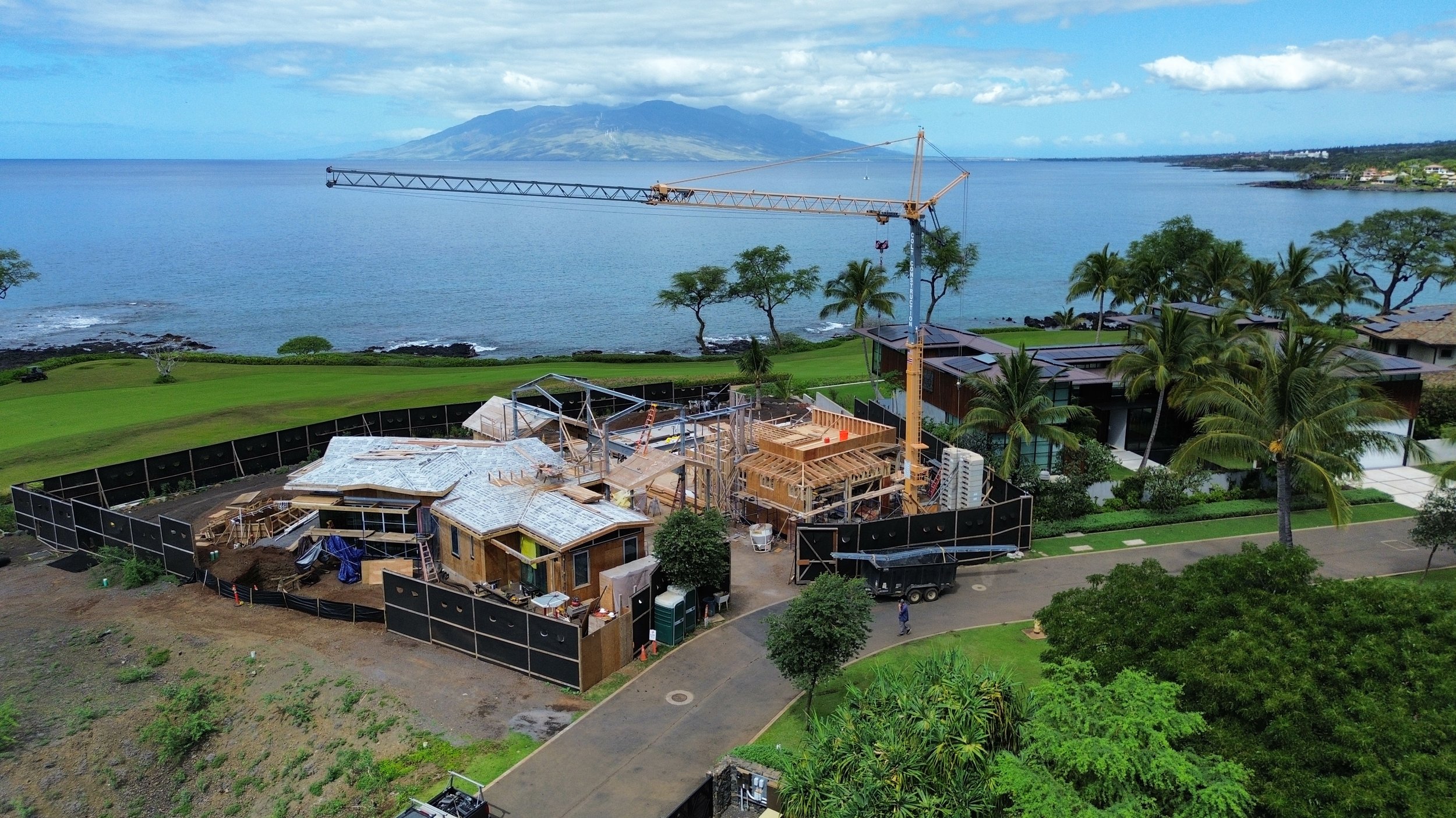 Construction site near the coastline with a crane, partly built wooden structures, lush greenery, palm trees, and mountains in the background.