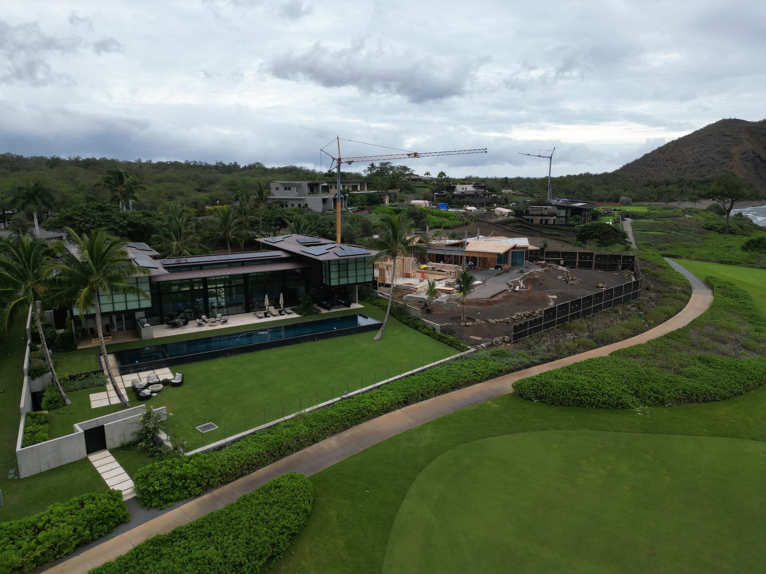 A modern house with a swimming pool, surrounded by green lawn and palm trees, under an overcast sky. There is ongoing construction on one side of the house, with construction cranes visible.