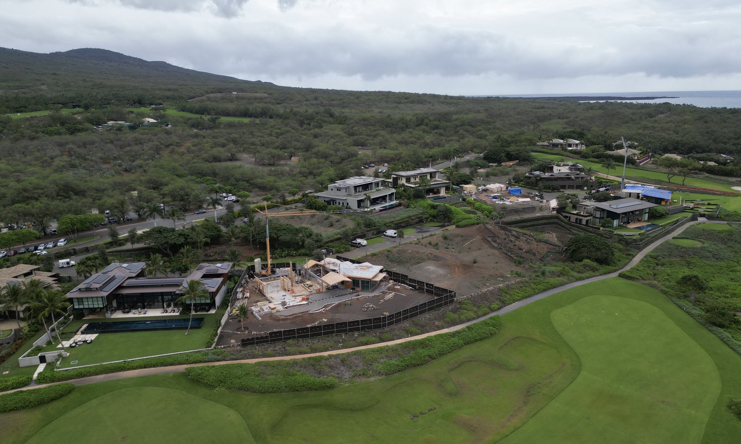 Aerial view of a residential area with a house under construction, neighboring houses, a golf course, and lush green landscape with hills in the background.