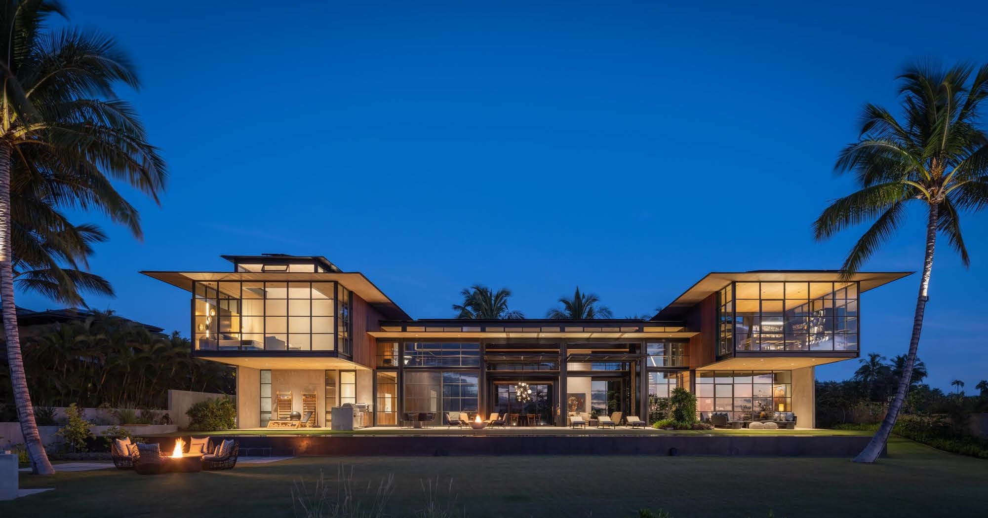 Modern two-story house with large glass windows, outdoor seating area, and palm trees, illuminated at dusk