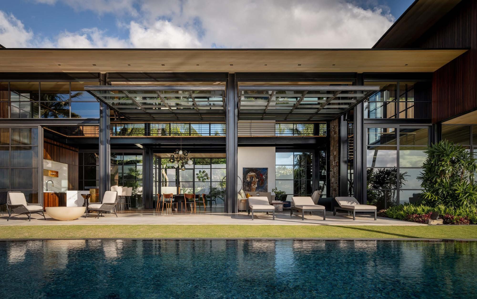 Modern house with large glass windows, a patio with lounge chairs and outdoor furniture, and a swimming pool in the foreground. The house features a combination of wood and metal elements with a lush garden setting.
