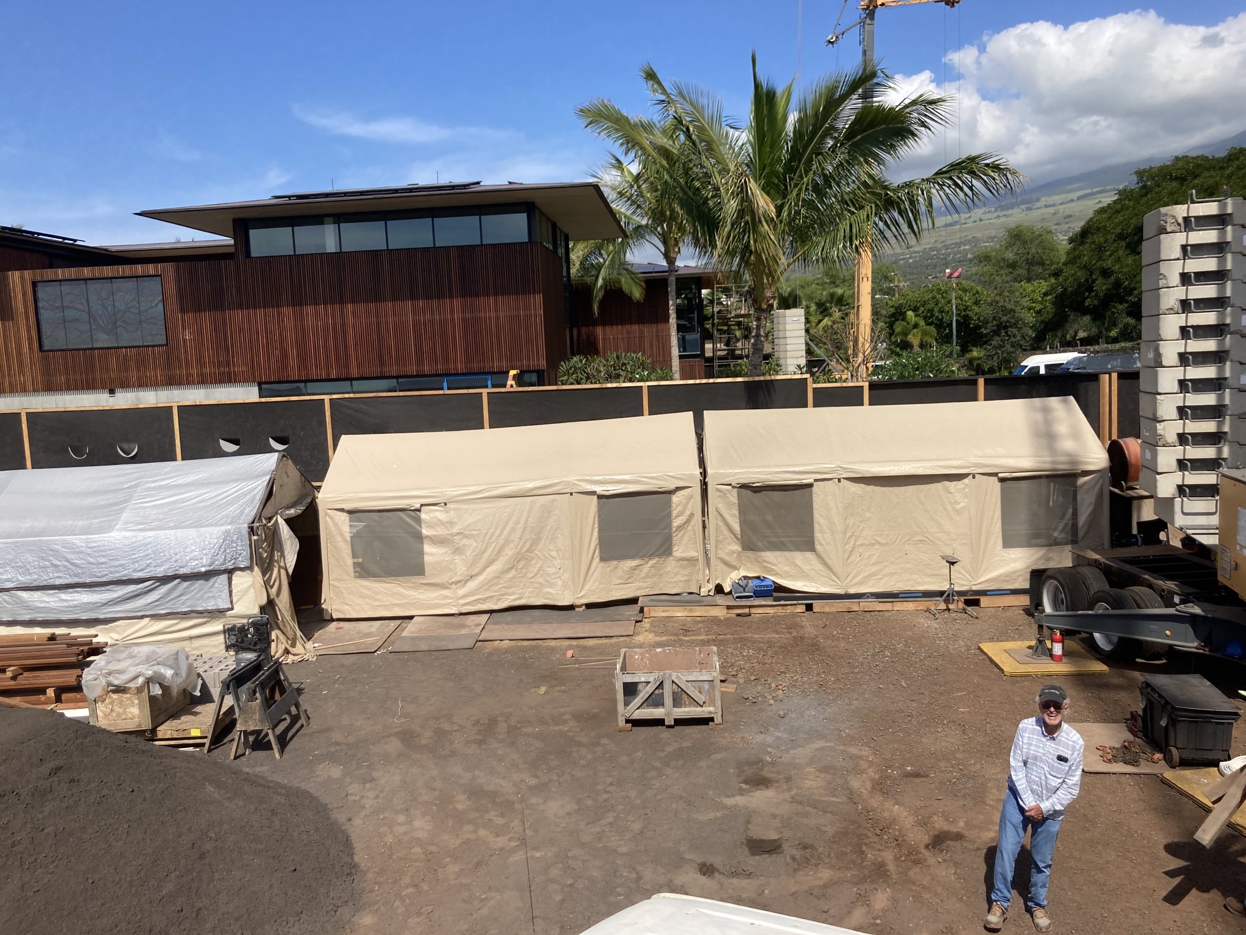 Construction site with large beige tents, a person standing in front, and a modern wooden house in the background, with palm trees and mountains.
