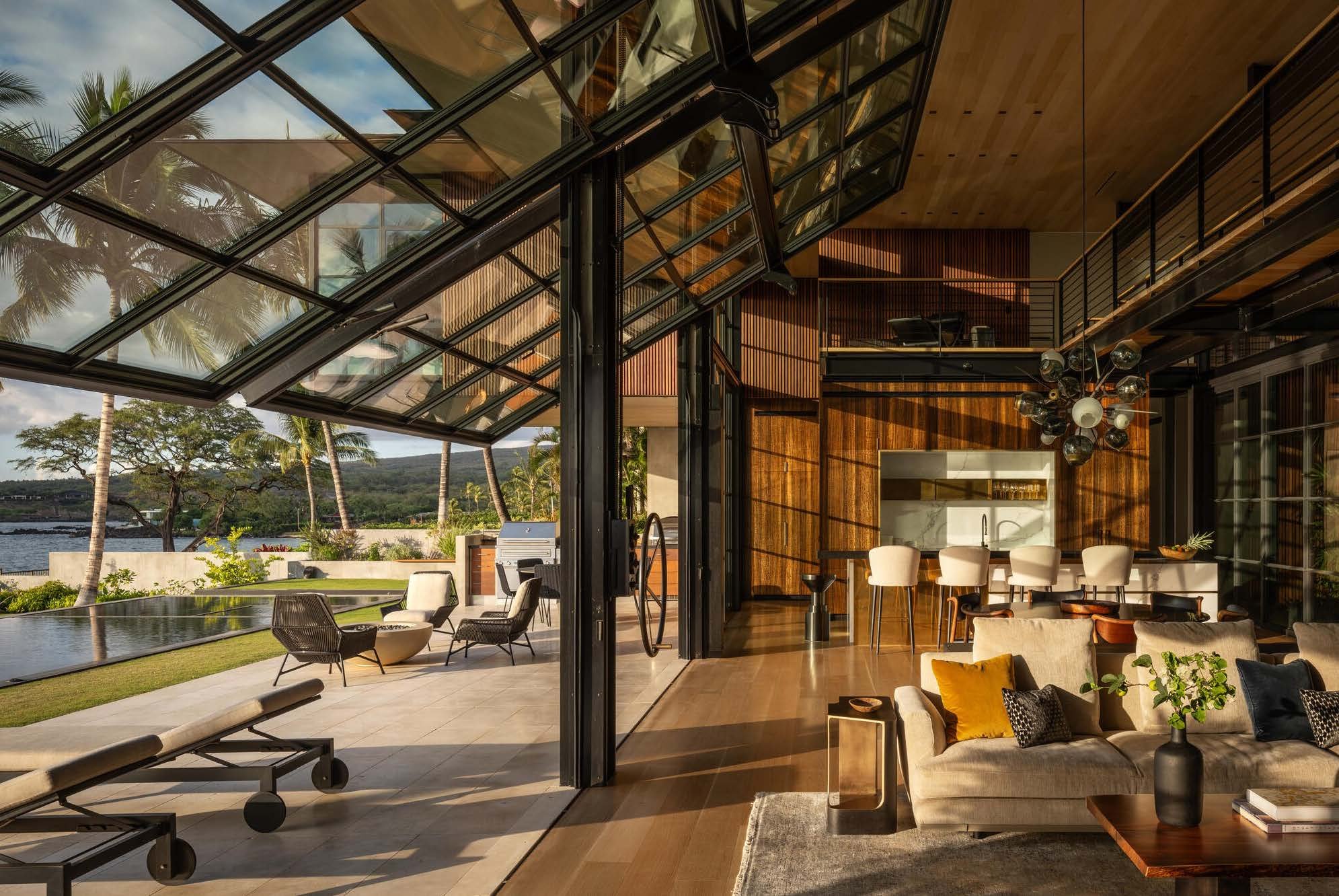 Modern open-concept living space with large glass doors and windows, wooden accents, a kitchen island with barstools, and an outdoor patio with lounge chairs, palm trees, and a body of water in the background.