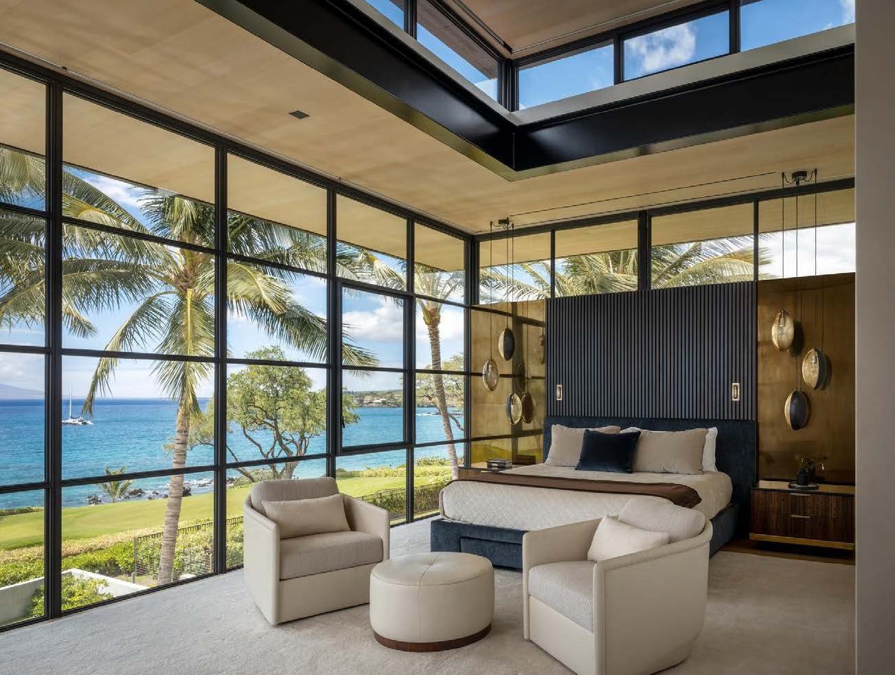 Modern bedroom with large floor-to-ceiling glass windows overlooking a tropical ocean view with palm trees, a lawn, and a boat in the water. The room features a large bed with pillows, hanging pendant lights on either side, a nightstand, and seating