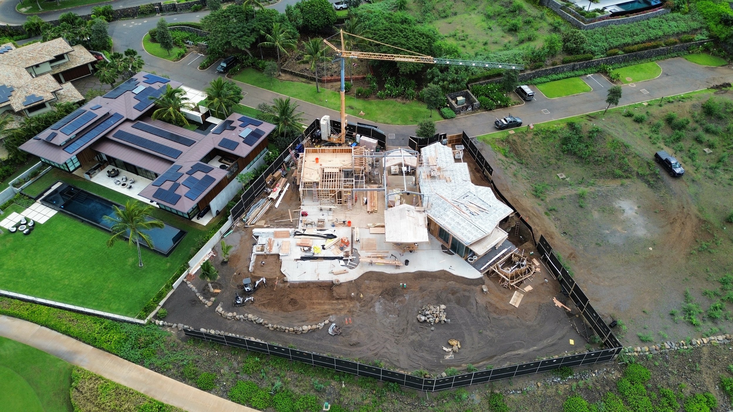Aerial view of a house under construction with a crane, surrounded by a black safety fence, neighboring finished houses with pools, green lawns, trees, and a parking lot.