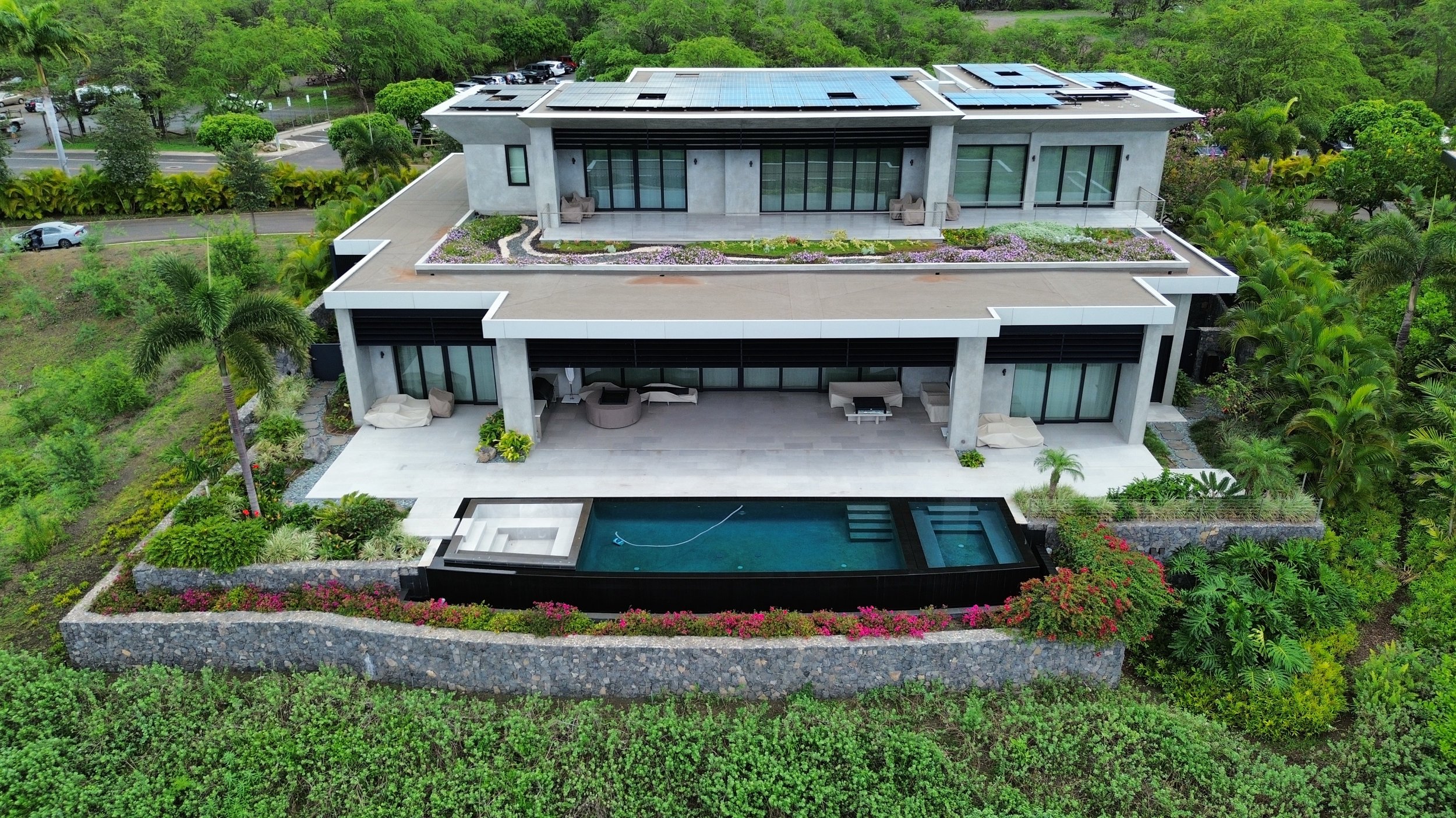 A modern multi-level house with a rooftop solar panel system, an infinity pool, and lush green landscape surrounding it.