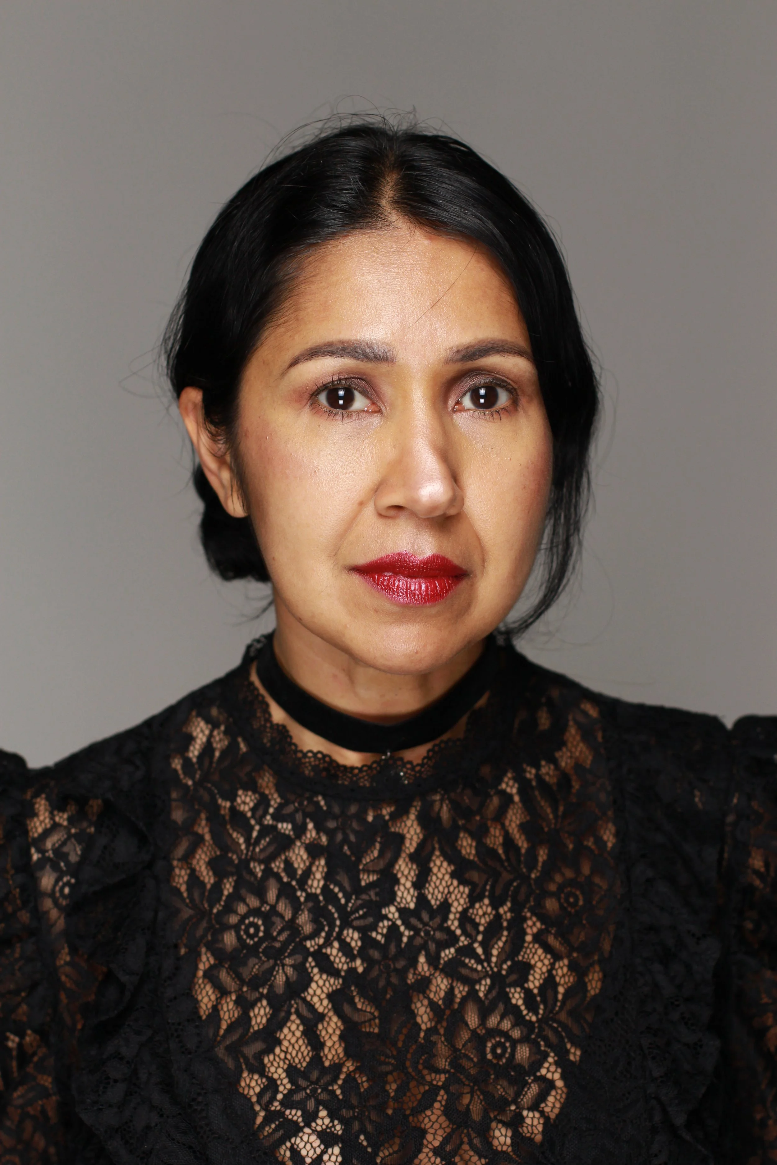 A woman with short black hair, wearing a black lace top, taking a selfie against a plain gray background.