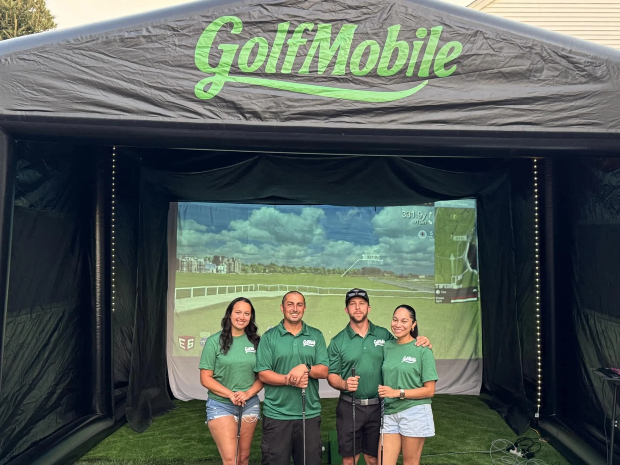 Four people in green golf shirts standing inside a golf simulation tent at GolfMobile, smiling and holding golf clubs, with a virtual golf course projected on a screen behind them.