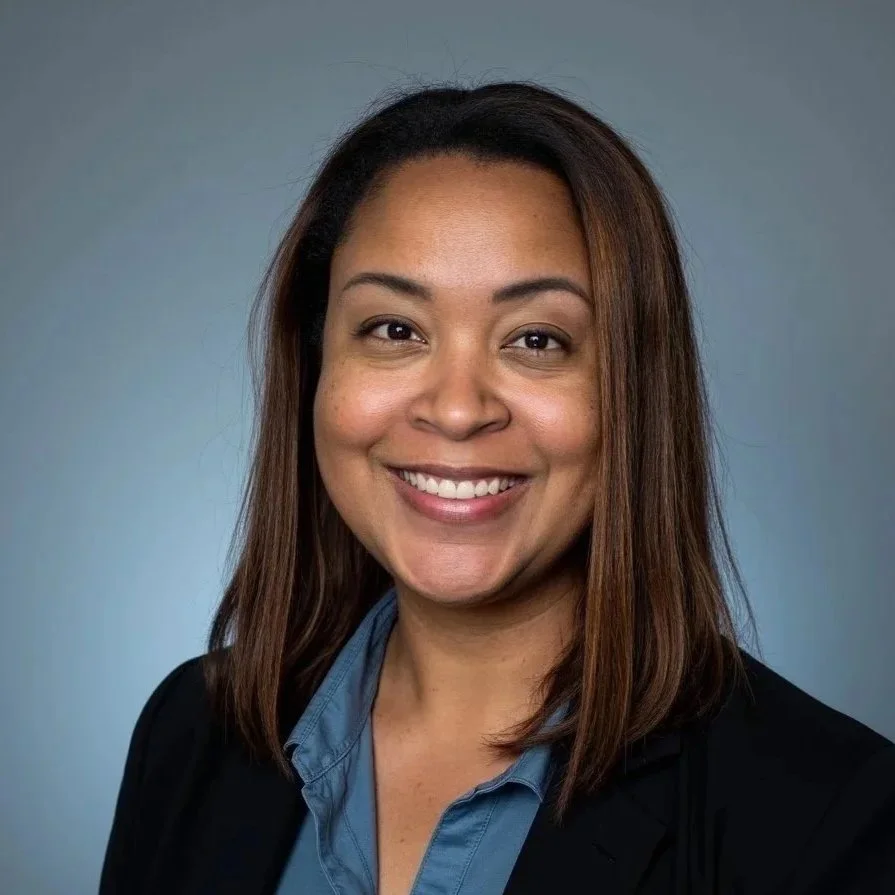 Professional woman with shoulder-length brown hair, wearing a dark blazer and blue shirt, smiling against a blue background.