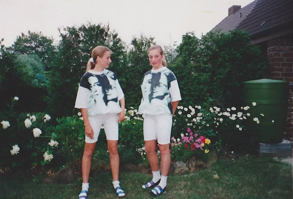 Two young teenage girls in matching white t-shirts, bike shorts, and sandals stand side by side in a lush German backyard garden, early 1990s.