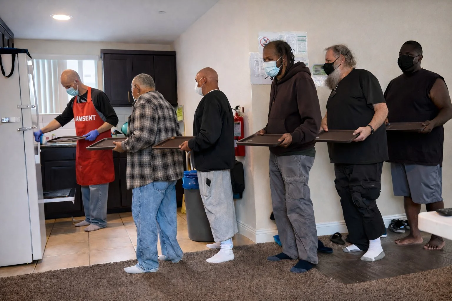 People standing in a line, wearing masks, waiting with trays in hand in a kitchen or food service area.