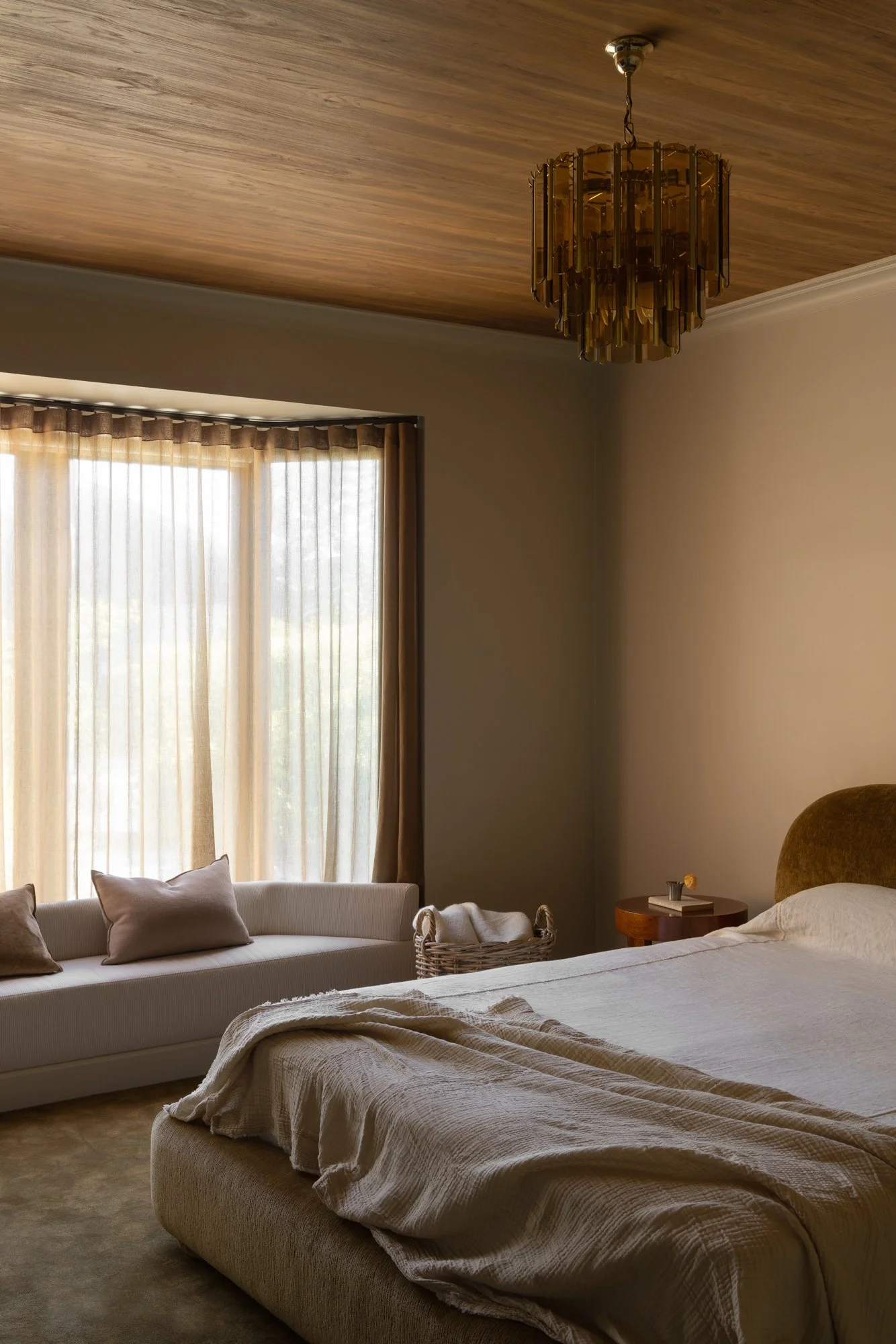 A cozy bedroom with a large bed covered in beige bedding, a small round wooden side table with a book and a cup, a basket beside the bed, large windows with beige curtains allowing natural light, a sofa with cushions near the window, and a decorative