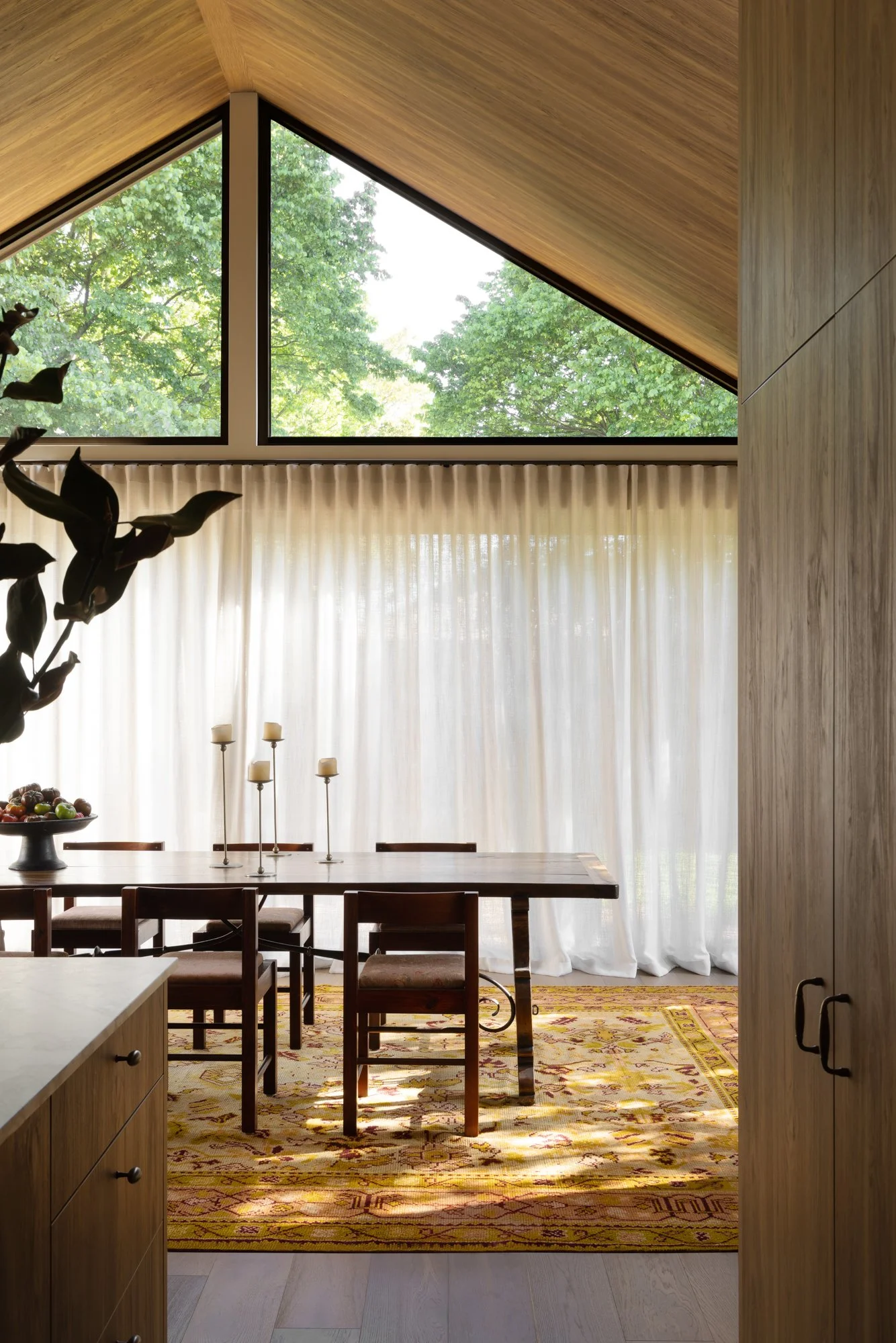 View of a dining room with a large wooden table, chairs, and a vintage patterned rug, seen through an open doorway. Large windows with sheer curtains and a sloped wooden ceiling are visible, and lush green trees are outside.