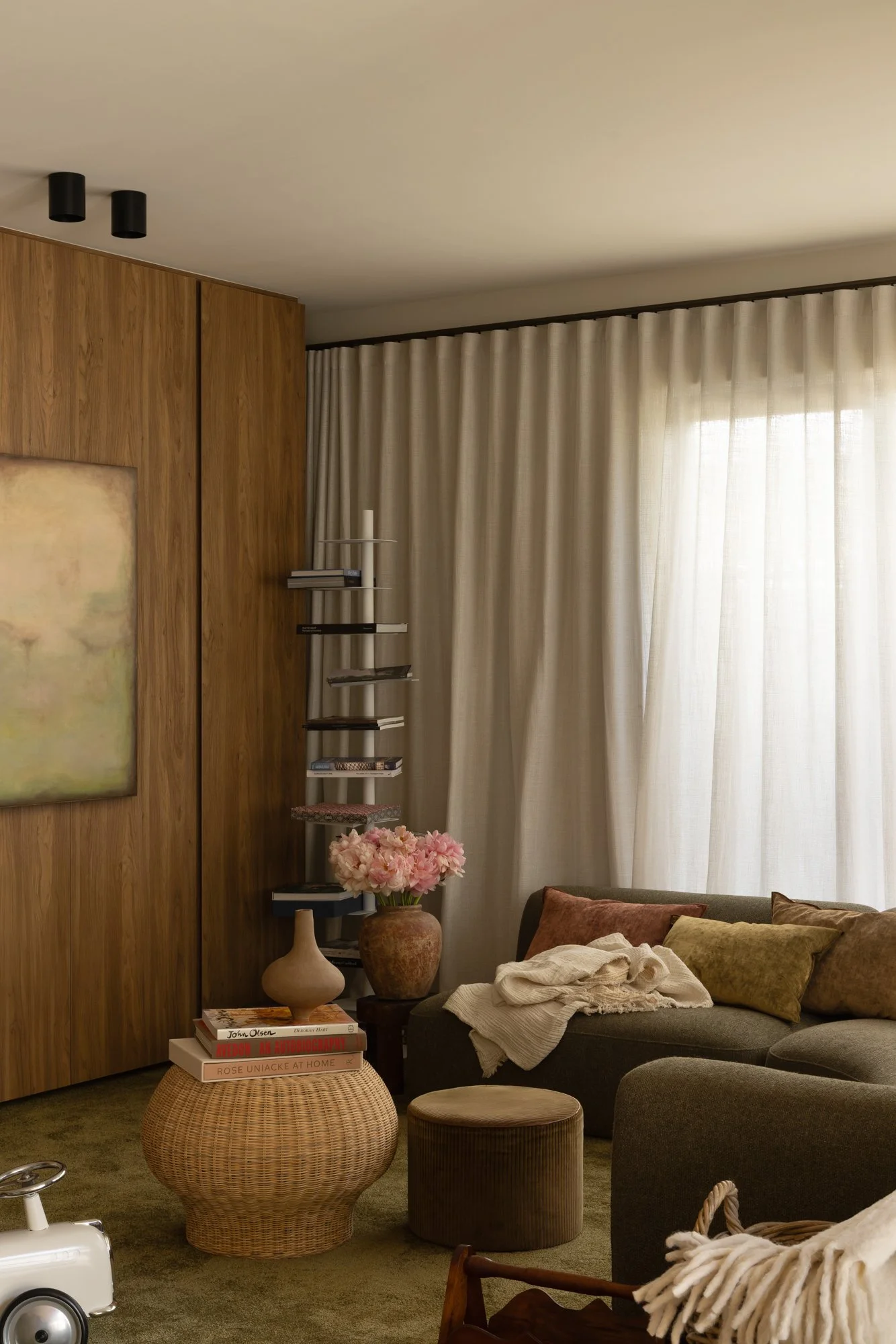Living room corner with a wooden wall, a bookcase, pink flowers on a vase, a sofa with pillows, and cream curtains.