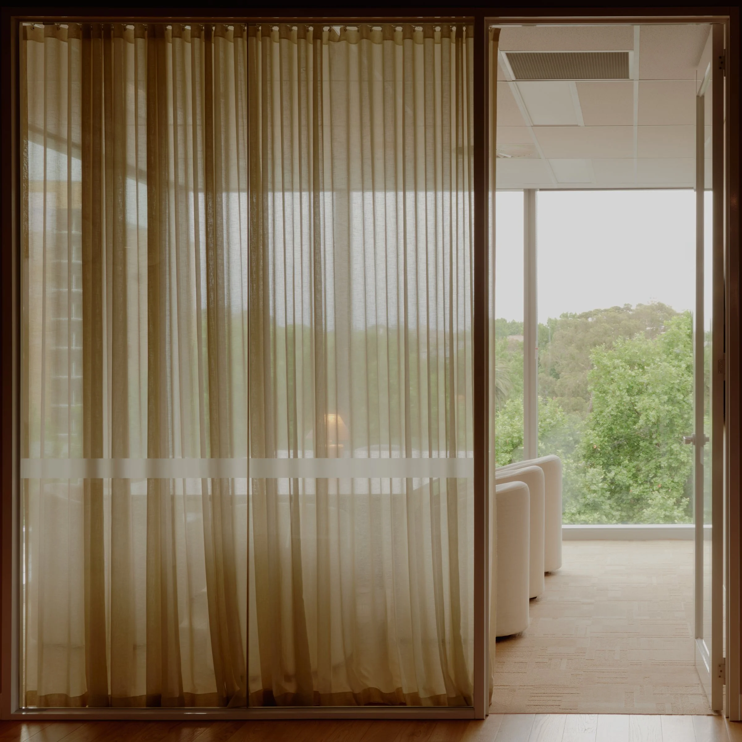 View through glass door with beige curtains to a seating area with white chairs and large windows showing trees outside.