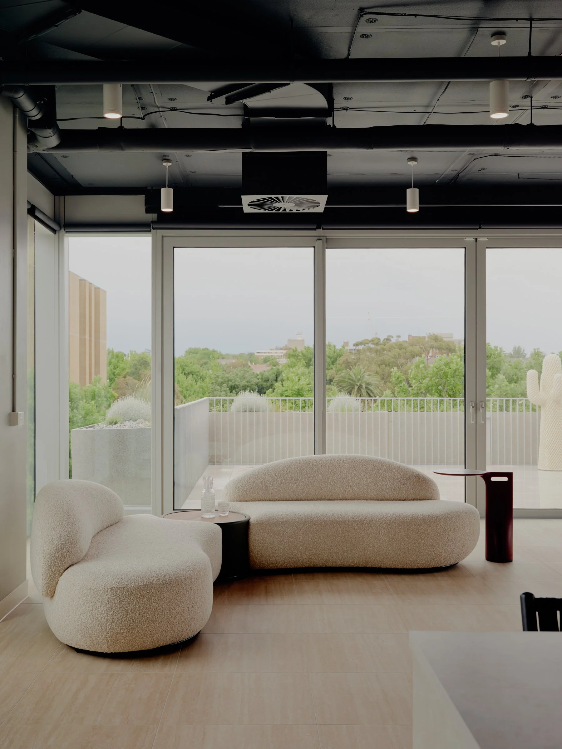 Modern indoor space with beige curved sofa and armchair, glass doors leading to a balcony with a view of trees and buildings, white and black ceiling fixtures, wooden floor.