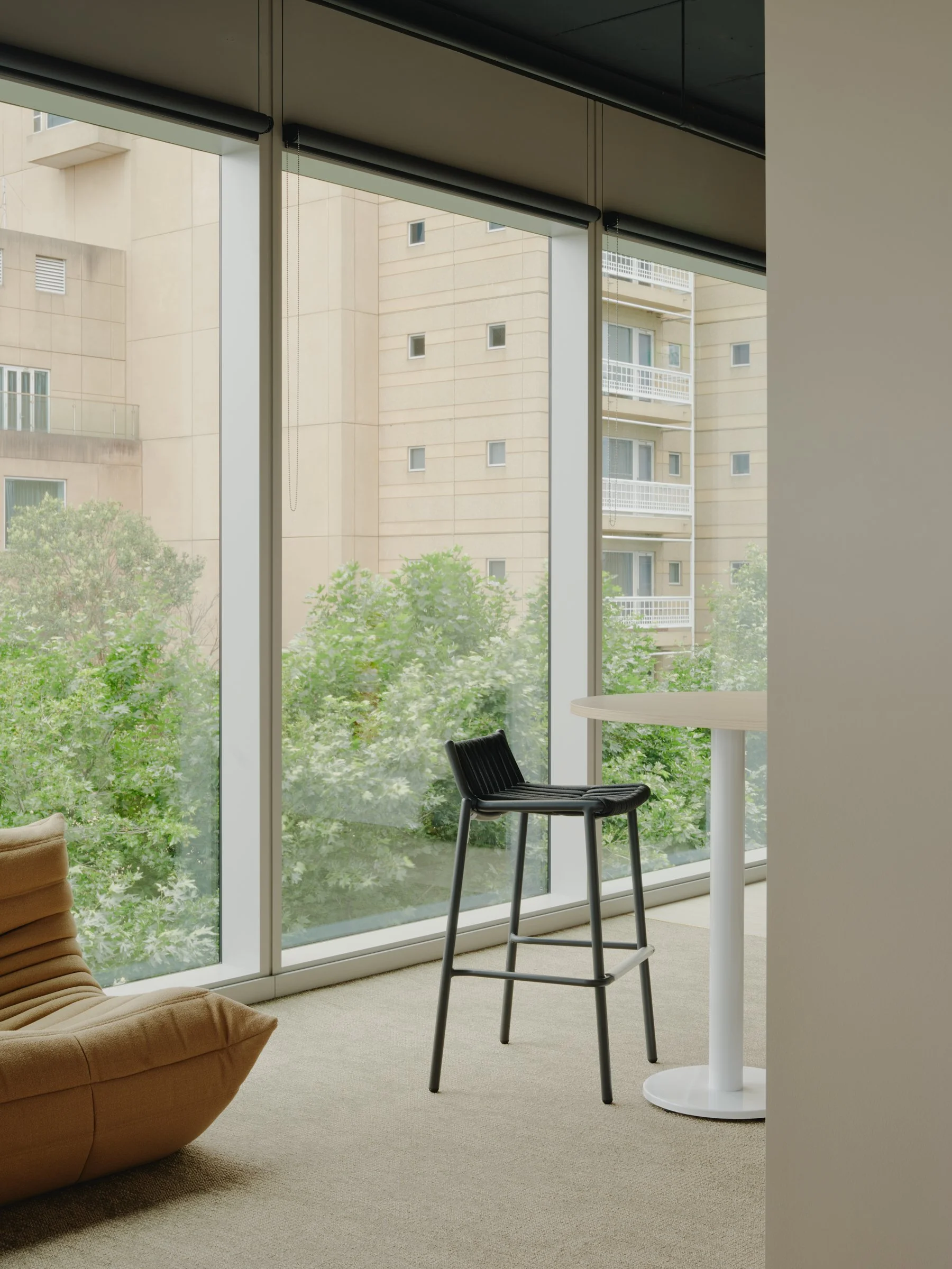 Interior of a modern room with large floor-to-ceiling windows facing a building and greenery outside, including a black chair and a small round white table.