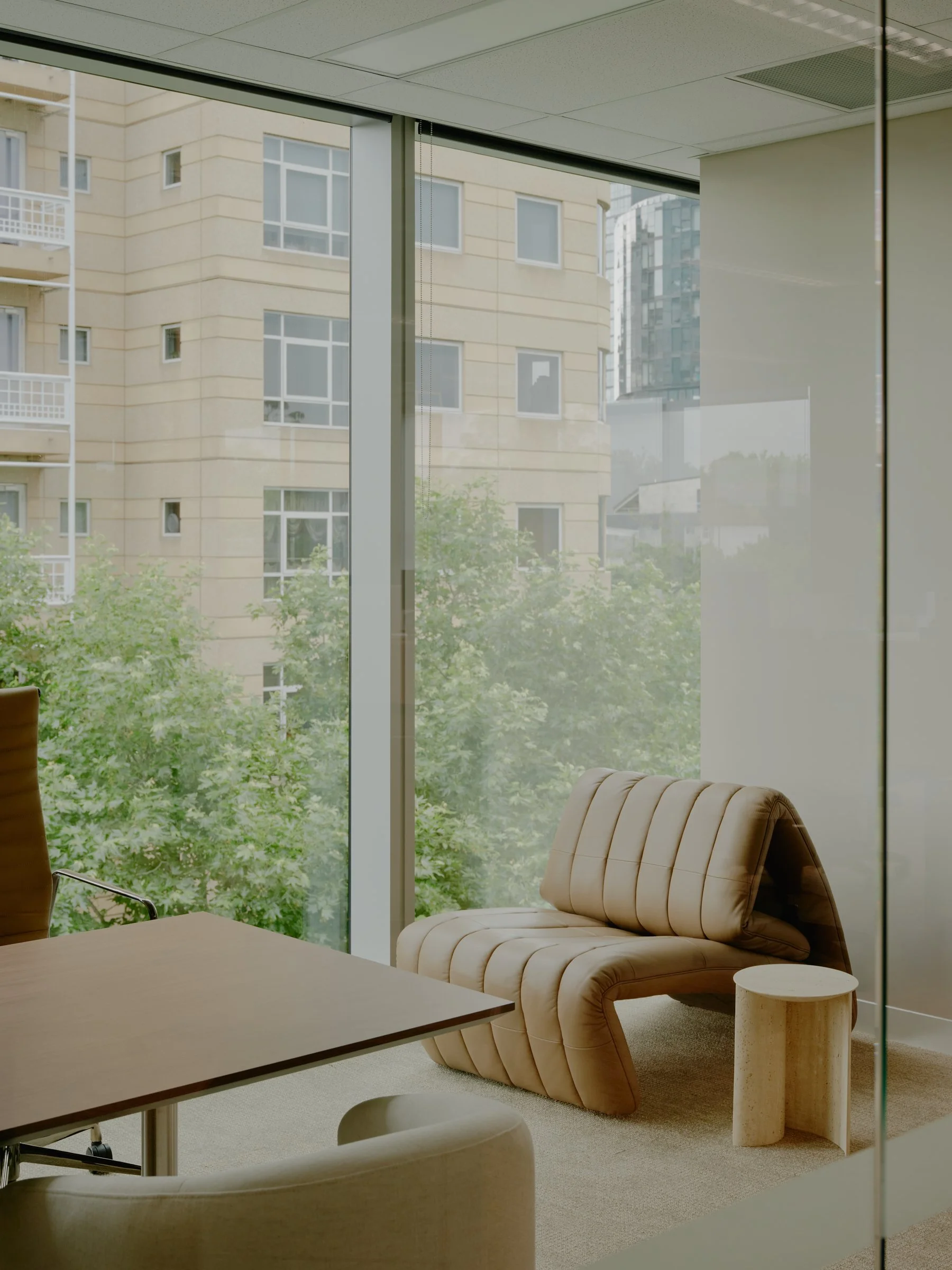 Modern office room with large glass window, beige ergonomic lounge chair, wooden side table, and part of office desk and chair.