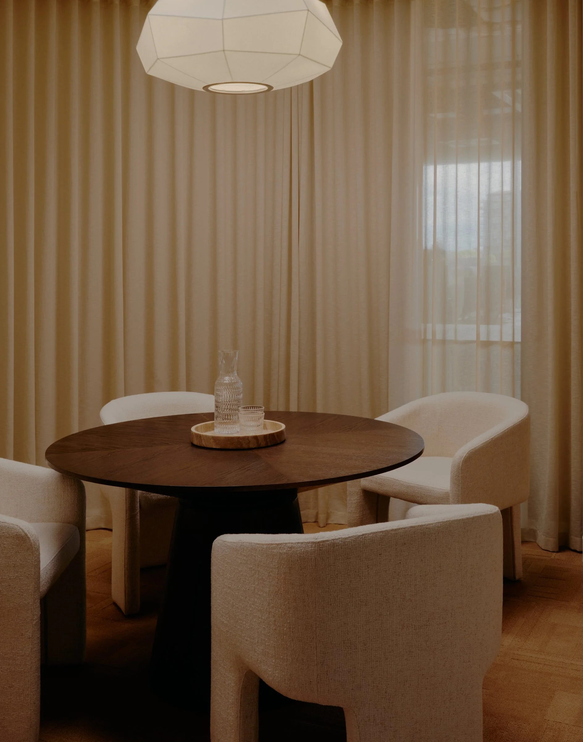 Dining room with a round wooden table, four beige upholstered chairs, a wooden tray with a glass and a pitcher, and beige curtains covering large windows, lit by a modern geometric pendant light.