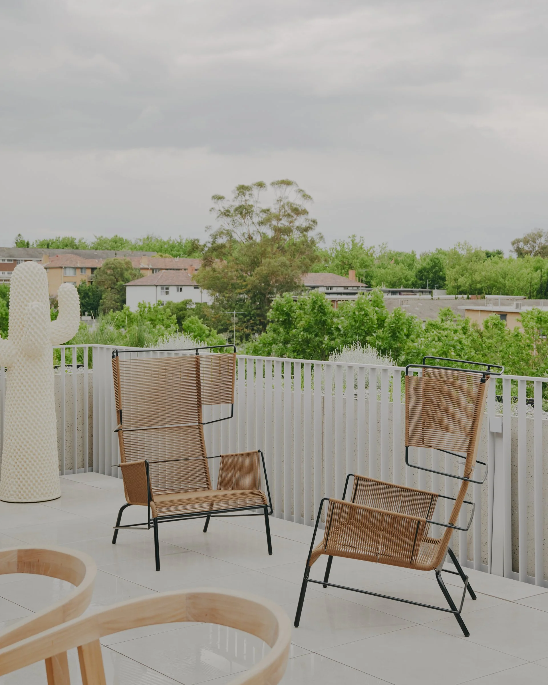 A balcony with two black metal and brown woven chairs, a large white cactus sculpture, a white railing, and a view of green trees and houses under a cloudy sky.