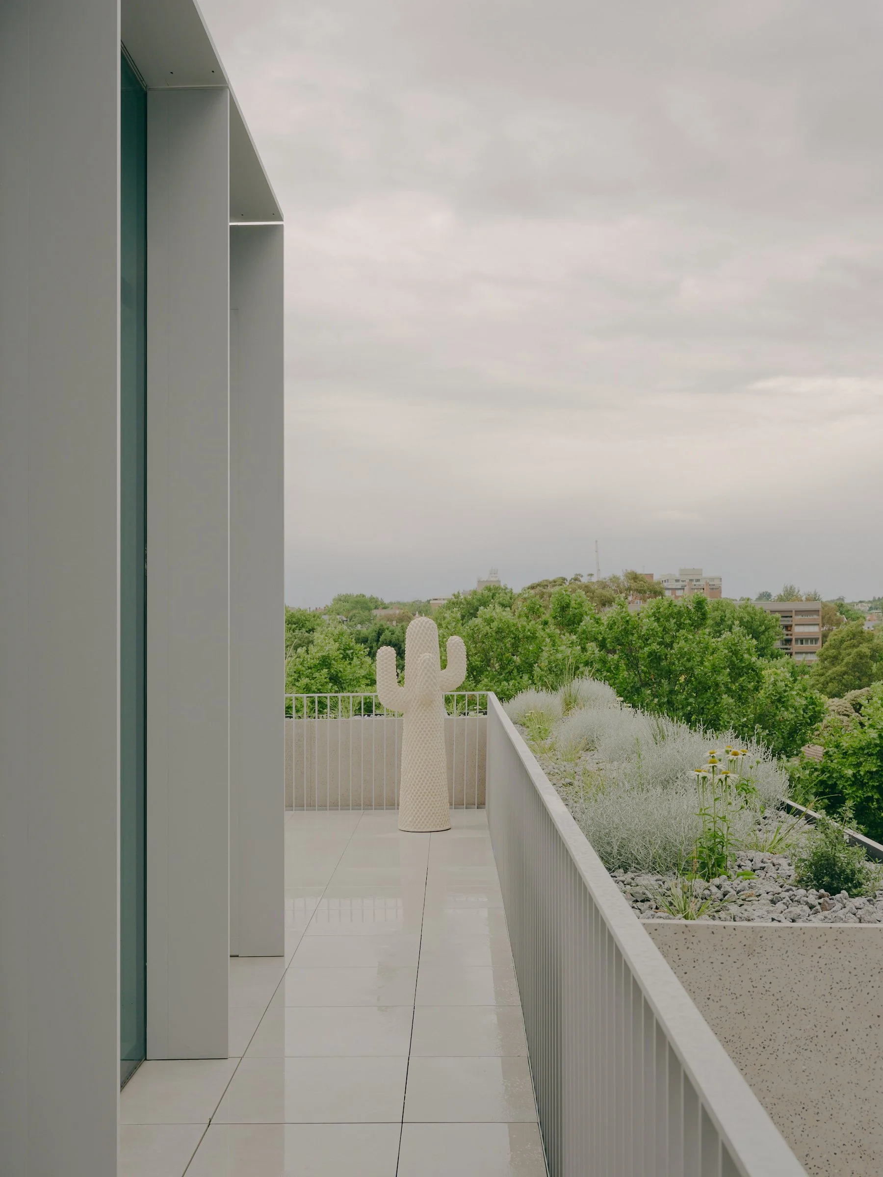Empty balcony with white tiled floor, white railing, and a white cactus-shaped sculpture, overlooking green trees and buildings under cloudy sky.