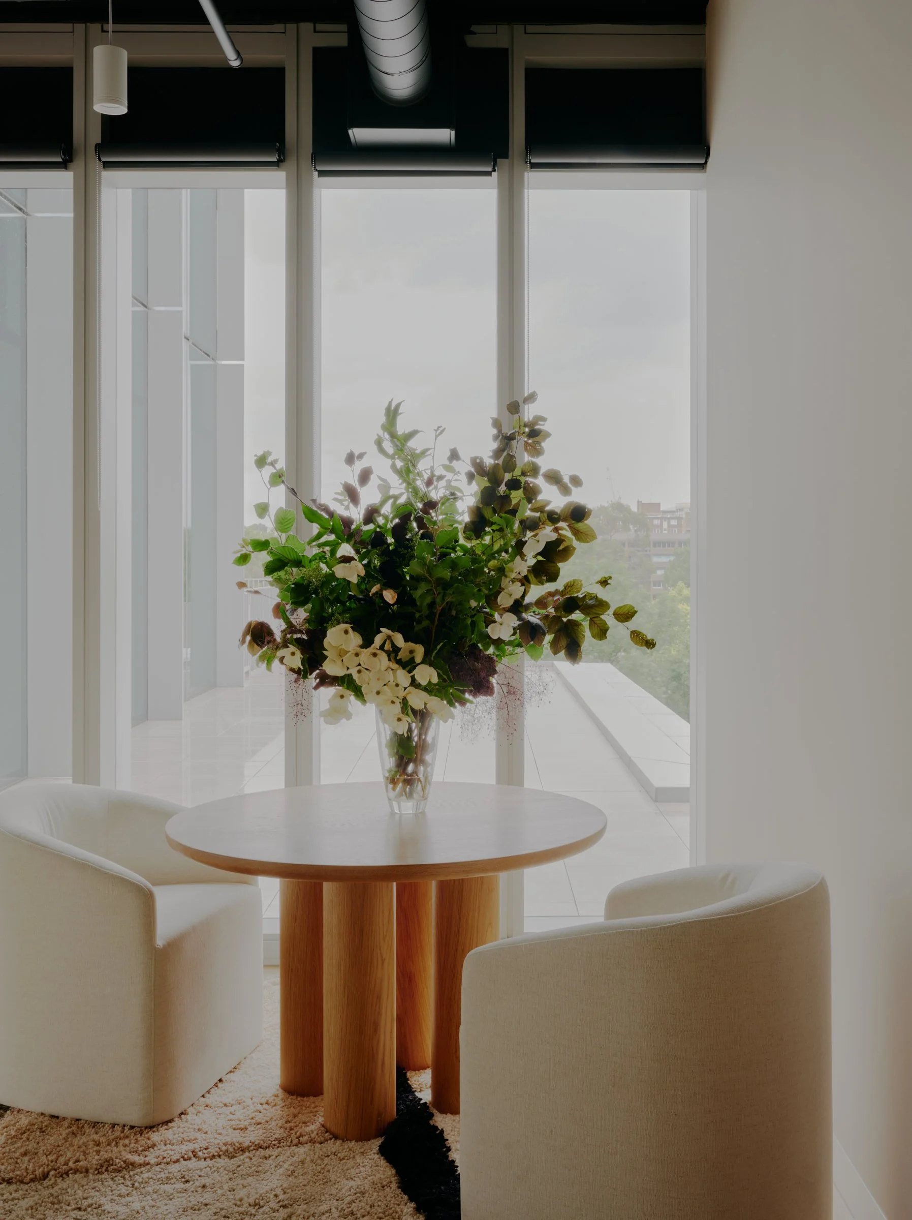 A round wooden table with a large flower arrangement in a glass vase, two white upholstered chairs, large glass windows with black shades, and a light-colored rug.