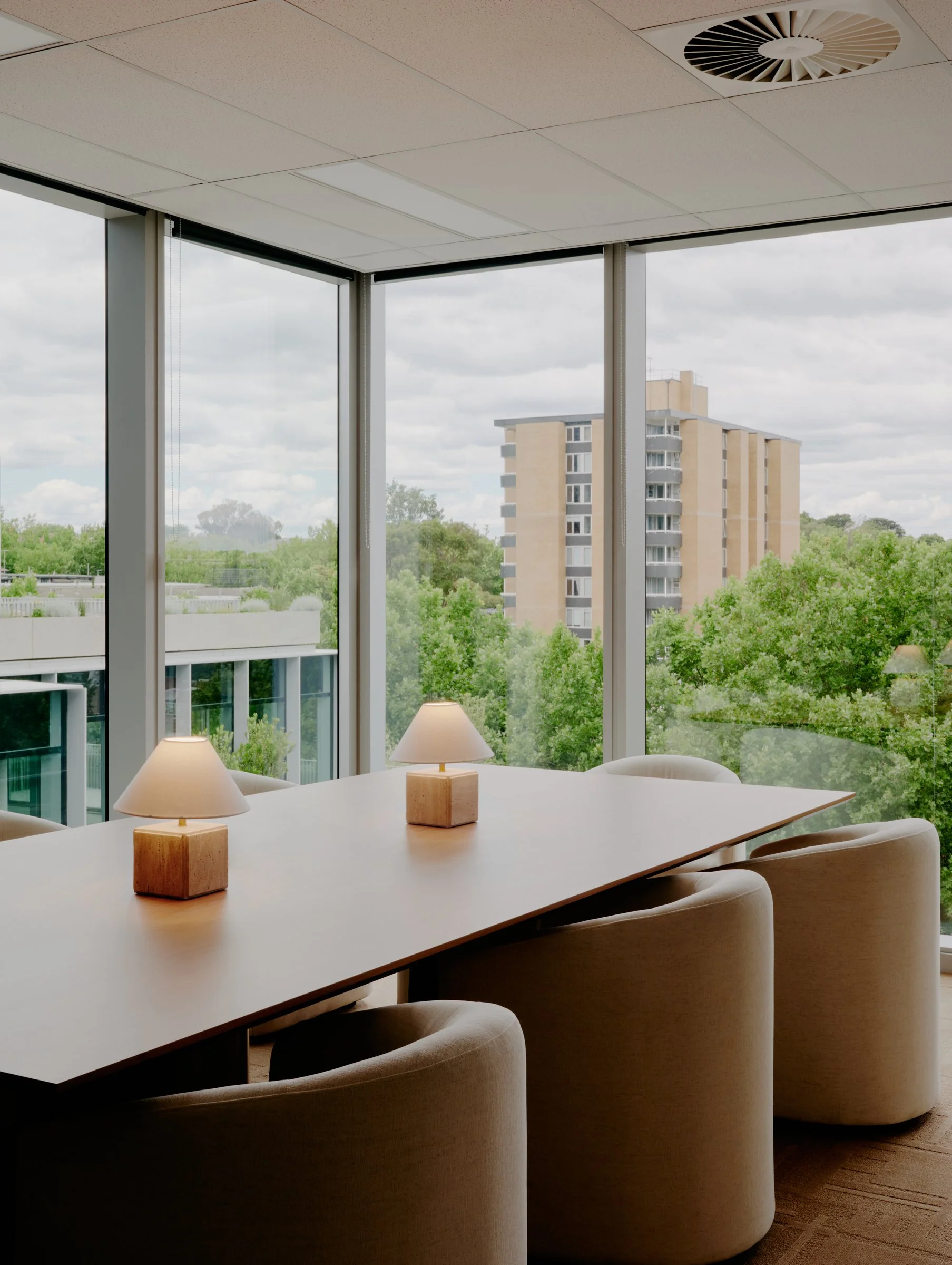 Interior view of a modern conference room with a large wooden table, surrounded by beige upholstered chairs, illuminated by two small table lamps, with floor-to-ceiling windows showing a cityscape with trees and a tall residential building.