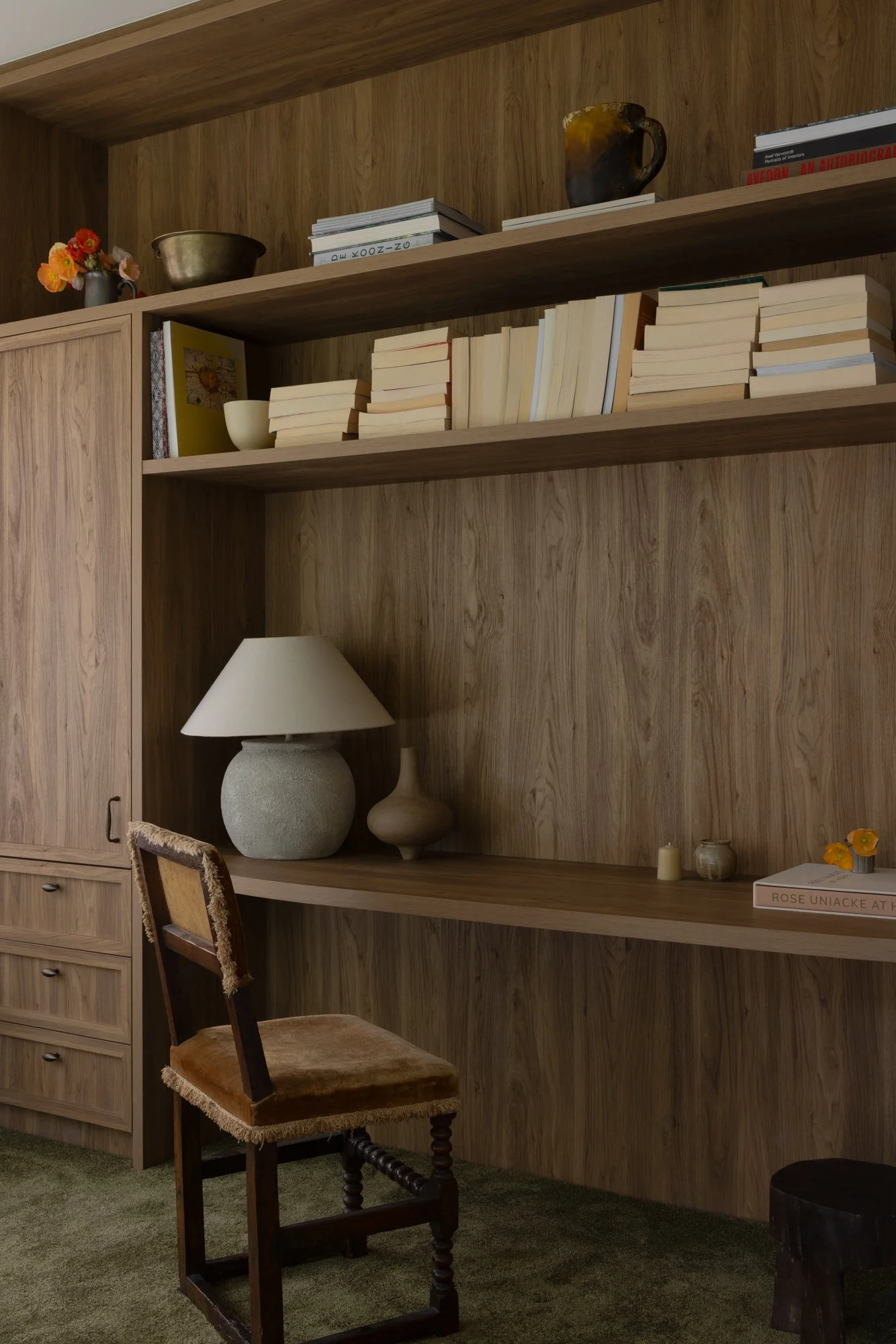 Wooden wall with shelves, decorated with books, ceramics, and vases; a wooden chair with a velvet seat in front, and a lamp on a shelf.