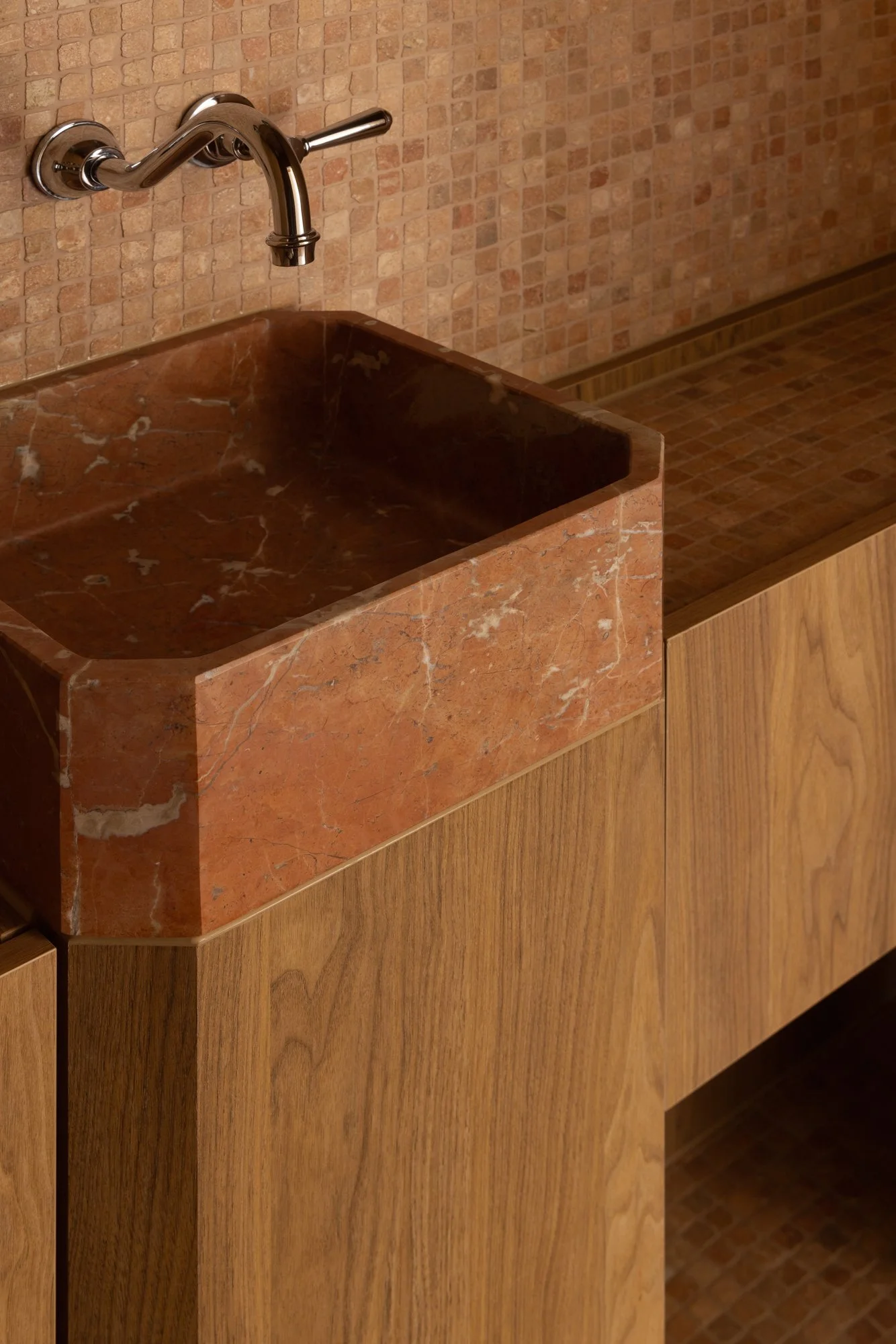 A wall-mounted metallic faucet above a built-in reddish-brown marble sink with a rectangular shape and rounded edges in a room with brick and wooden wall and paneling.