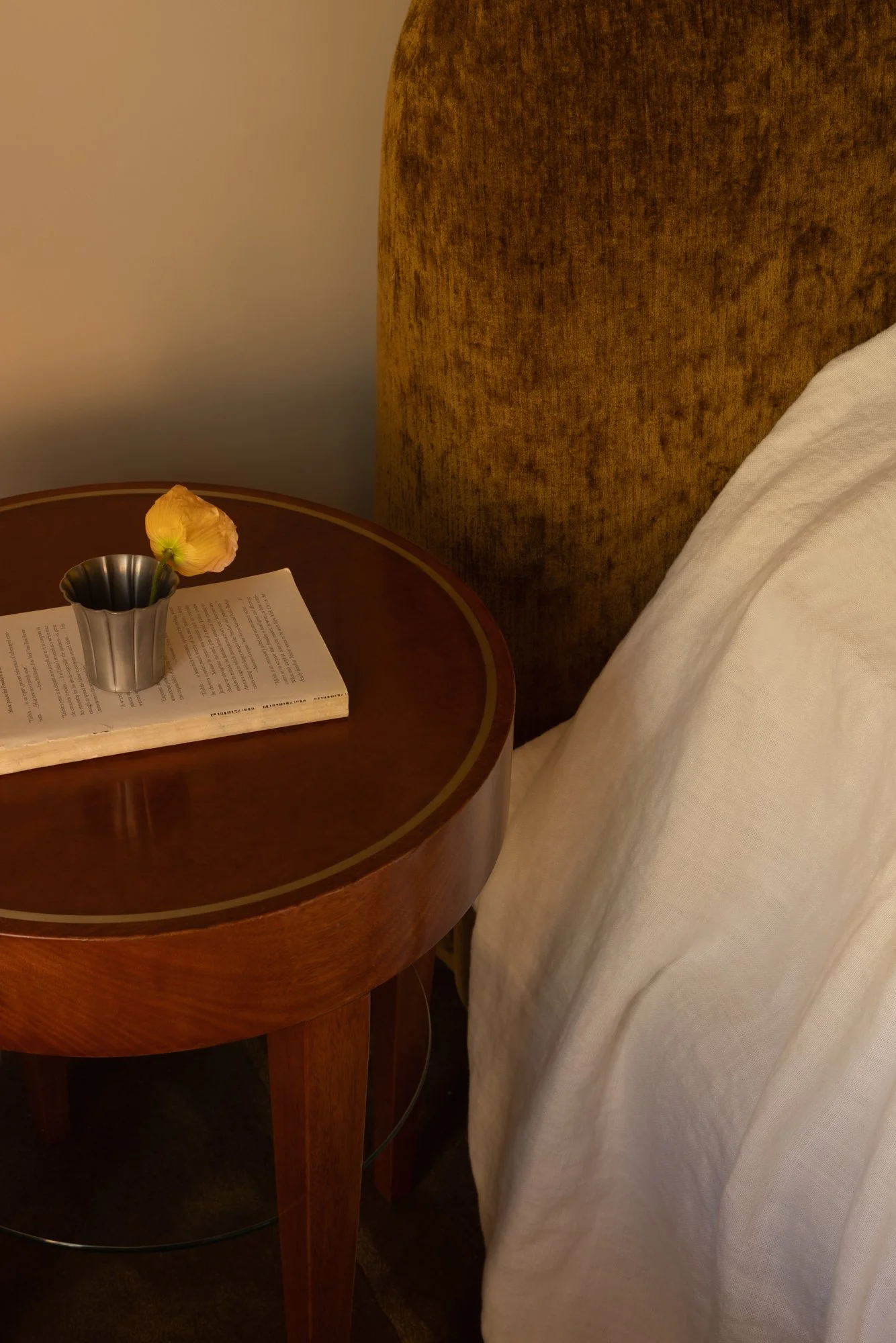 Close-up of a wooden nightstand with a book, a vase with a single flower, and a bed with white sheets and a mustard-colored headboard.