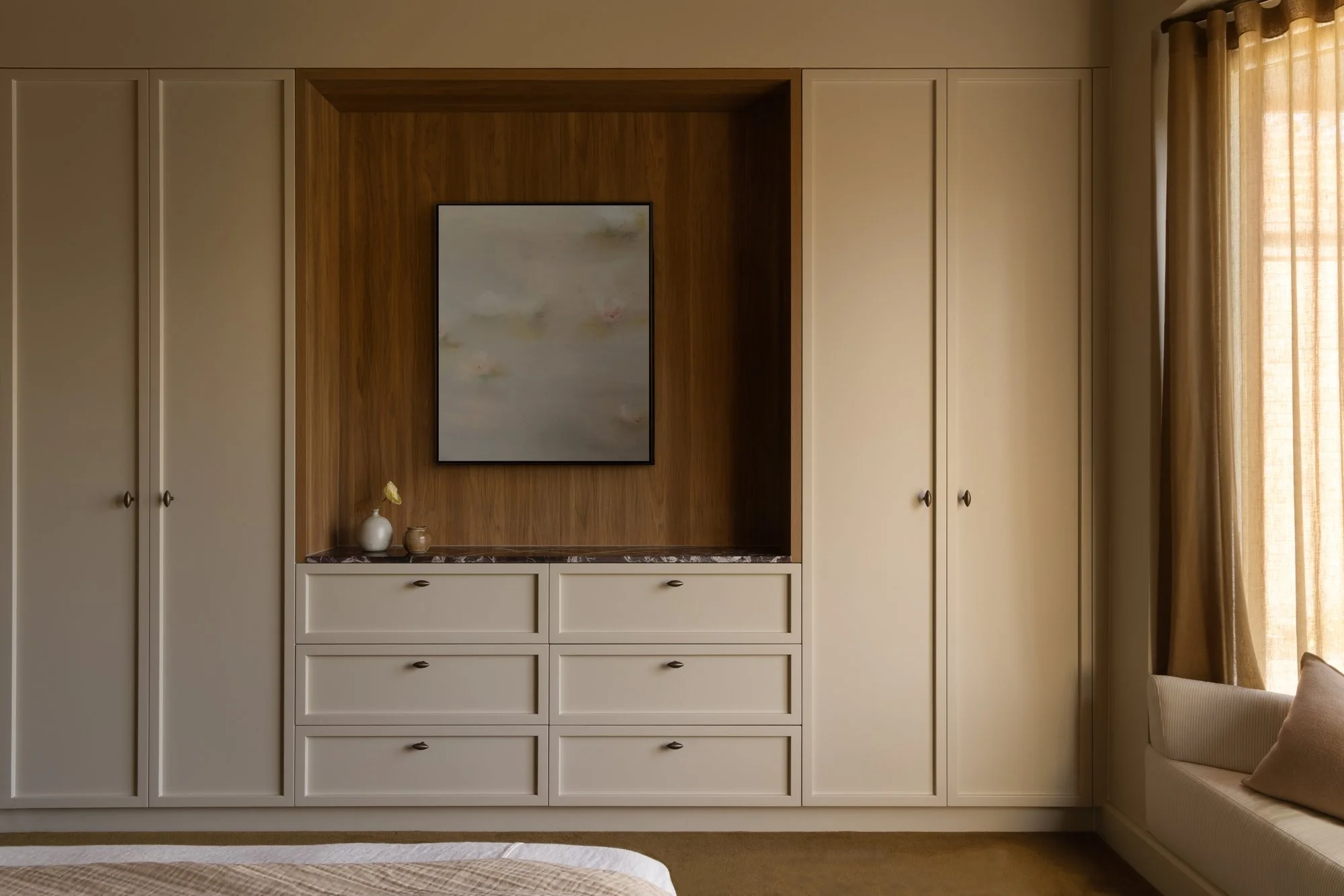 Built-in beige wardrobe with wood accents in a bedroom, featuring a framed abstract painting, a white vase with a yellow flower, and a small brown decorative object, next to a window with beige curtains.