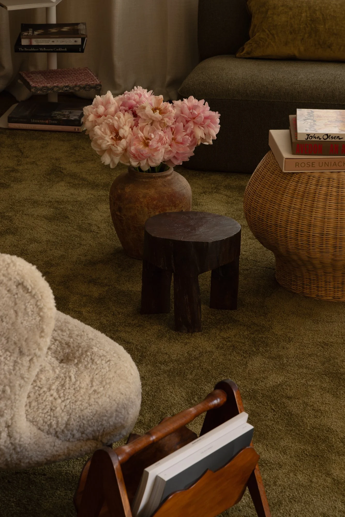 Living room with a large bouquet of pink flowers in a rustic vase on a dark wooden stool, a beige shaggy armchair in the foreground, a sofa with brown and yellow cushions, and a side table with several books and magazines in the background.