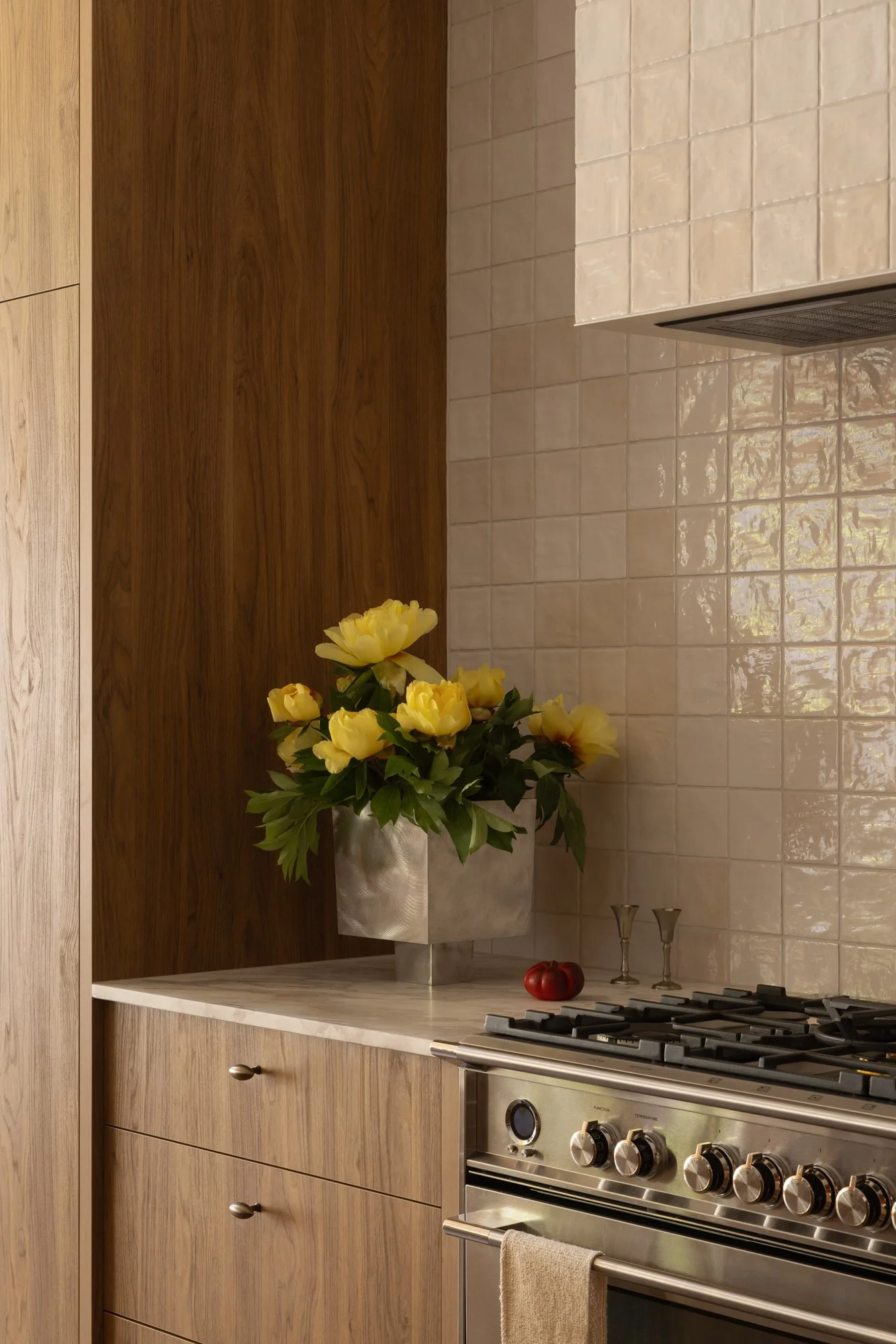 A kitchen with wood-paneled cabinets, a white marble countertop, and a stainless steel stove. There is a large silver vase with yellow flowers on the counter, a single red tomato, and two wine glasses. The backsplash wall is tiled with beige tiles.
