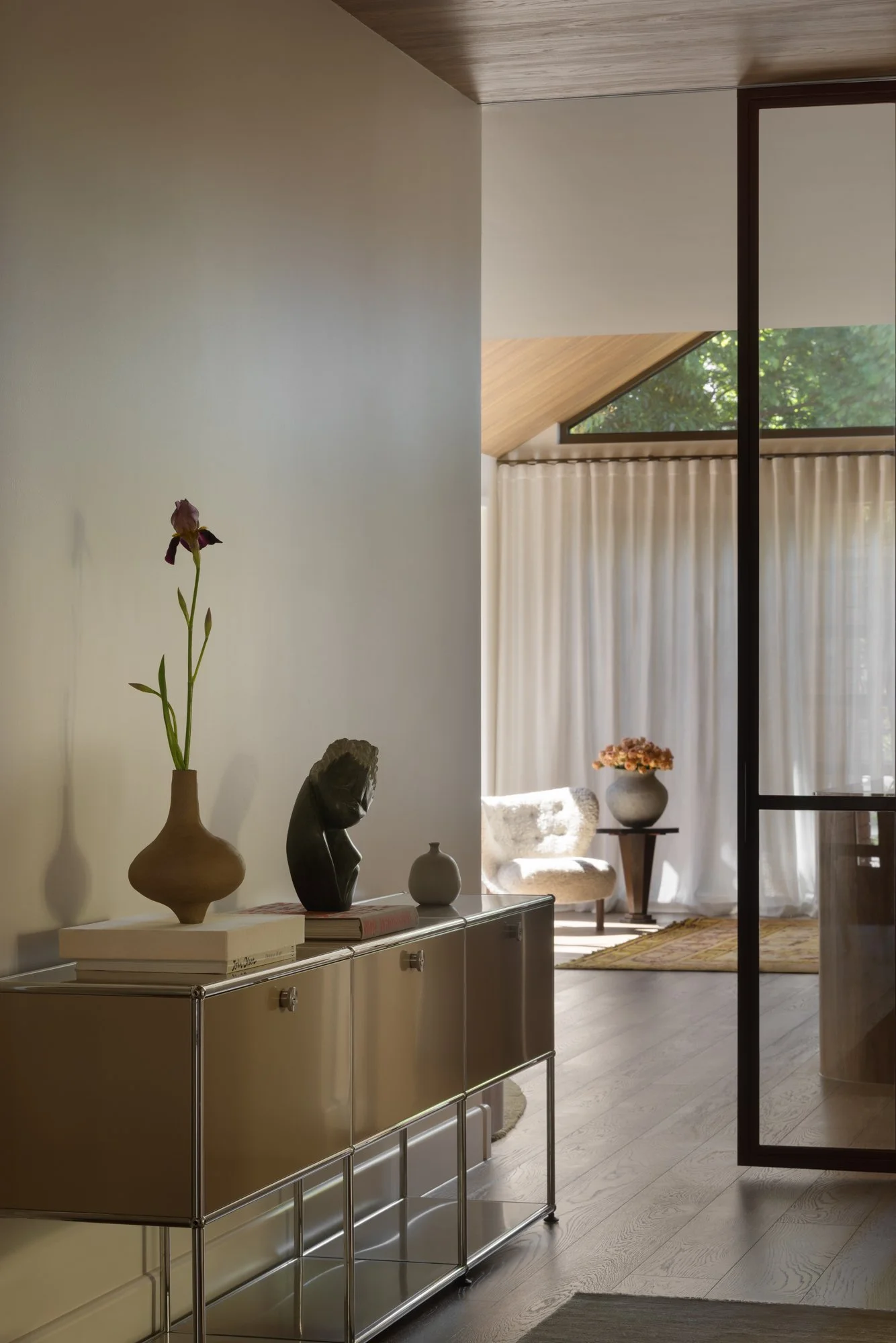 A modern interior room with a beige sideboard decorated with vases and sculptures, a small table with a flower arrangement, a cozy armchair, and a sliding glass door leading to a room with large windows, curtains, and a view of trees outside.