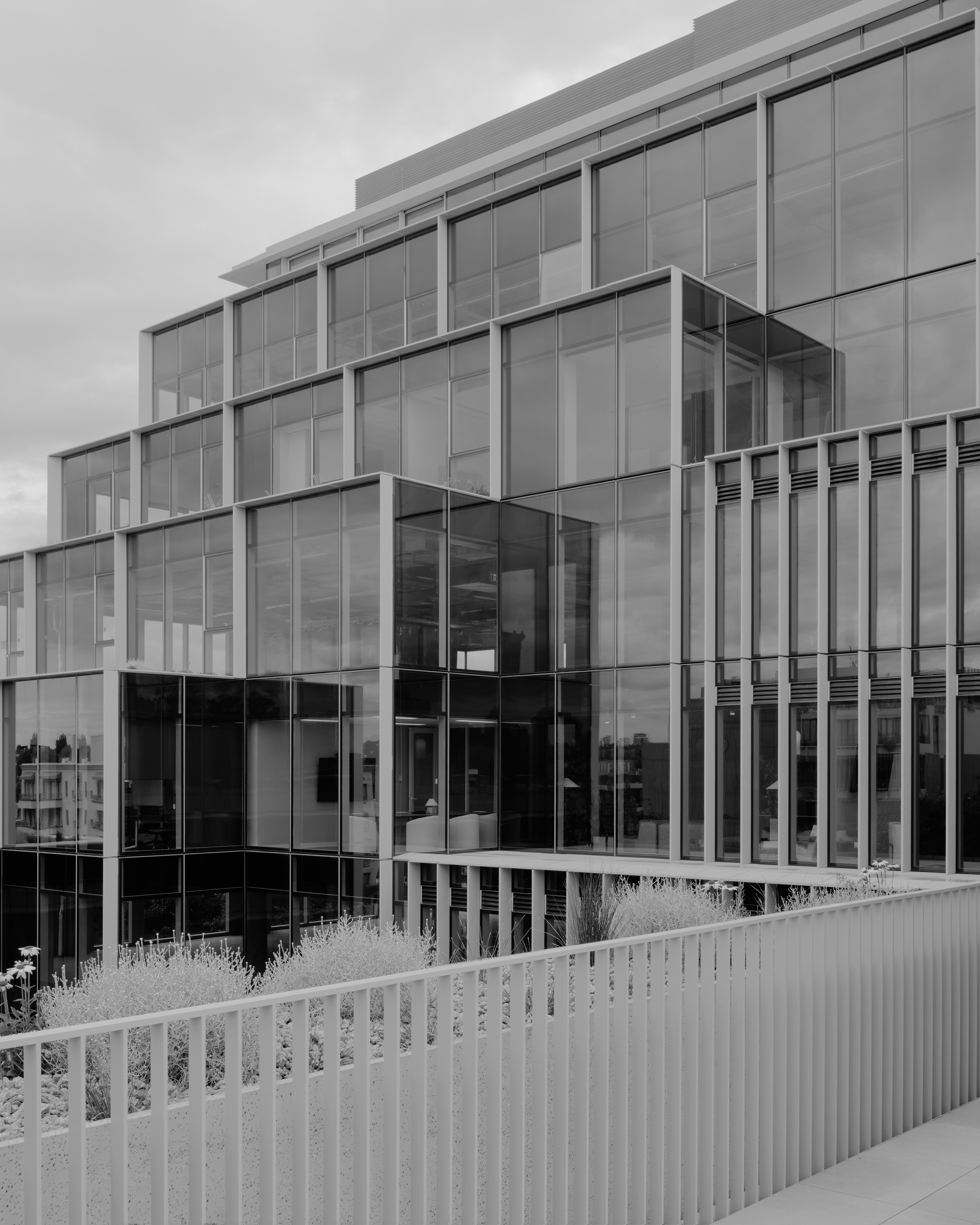 Modern glass building with multiple floors, featuring large windows and a balcony. The structure has a sleek, contemporary design with clean lines and geometric shapes. There is a white picket fence and some landscaping in the foreground.